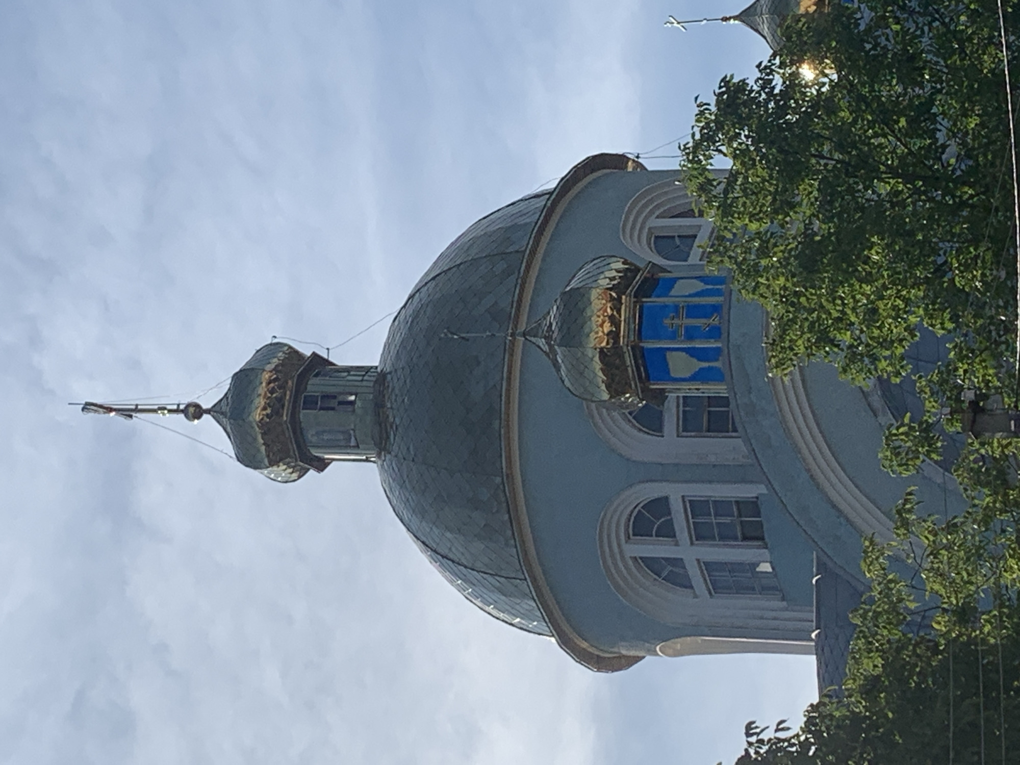 The image shows the central dome and a smaller dome of the Transfiguration Cathedral in Kolomyia, Ukraine. The main dome is large and covered in greenish-grey shingles, with a band of gold trim near its base. It is supported by a light blue cylindrical structure that features arched windows with white trim. A smaller, golden dome is positioned to the right of the main dome, featuring blue panes with a cross motif. Both domes are topped with crosses. The upper part of a large tree with green leaves obscures the lower portion of the cathedral. The sky above is partly cloudy with a bright, clear blue sky visible in some areas. The lighting suggests it is daytime, possibly late afternoon due to the angle of the sun.