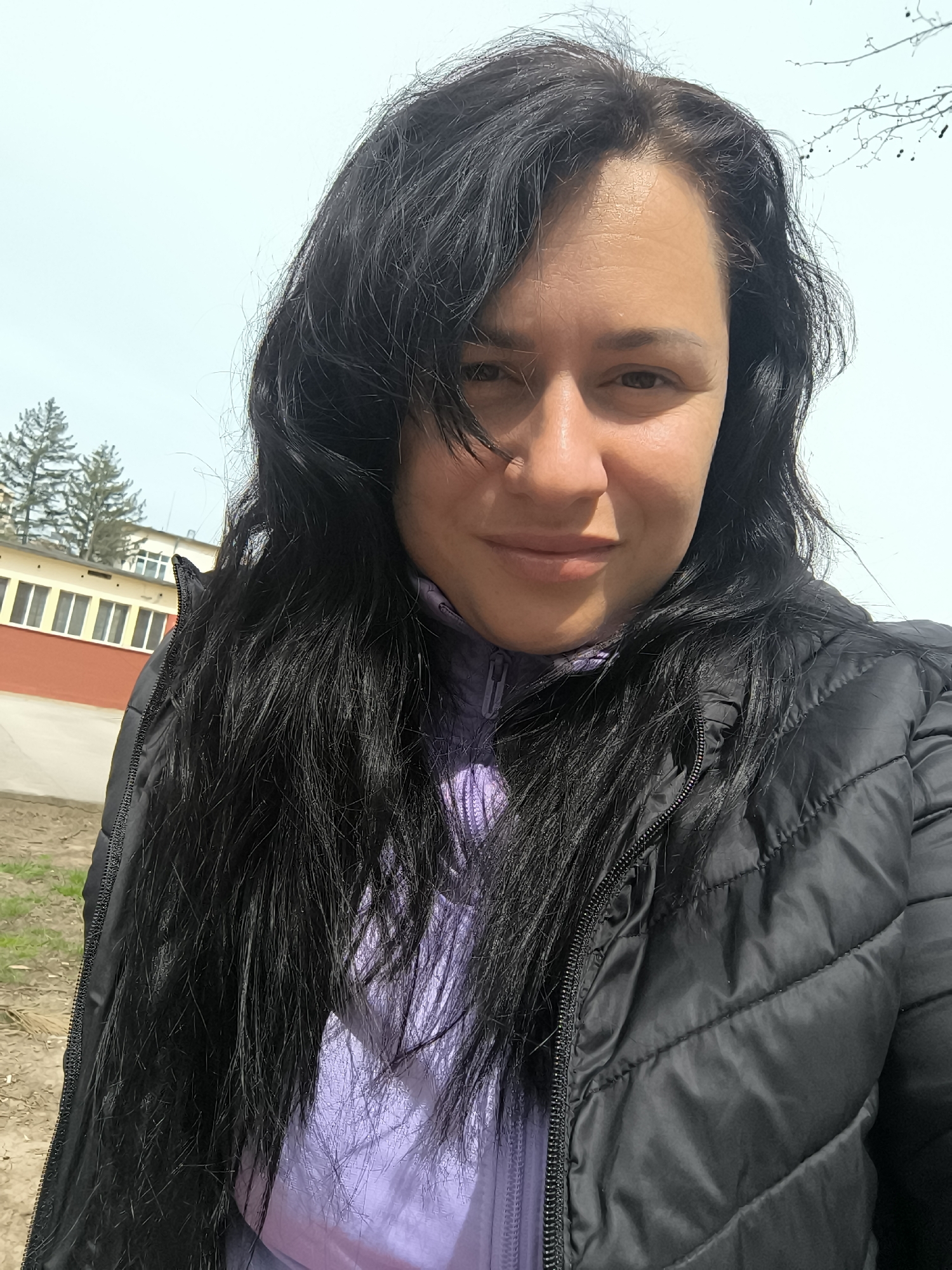 A woman with dark hair and fair skin is facing the camera. She is wearing a black puffer jacket over a light purple top. In the background, there is a red and yellow building with windows, and pine trees are visible behind it. The sky is light blue and overcast. The location is Pleven, Bulgaria.