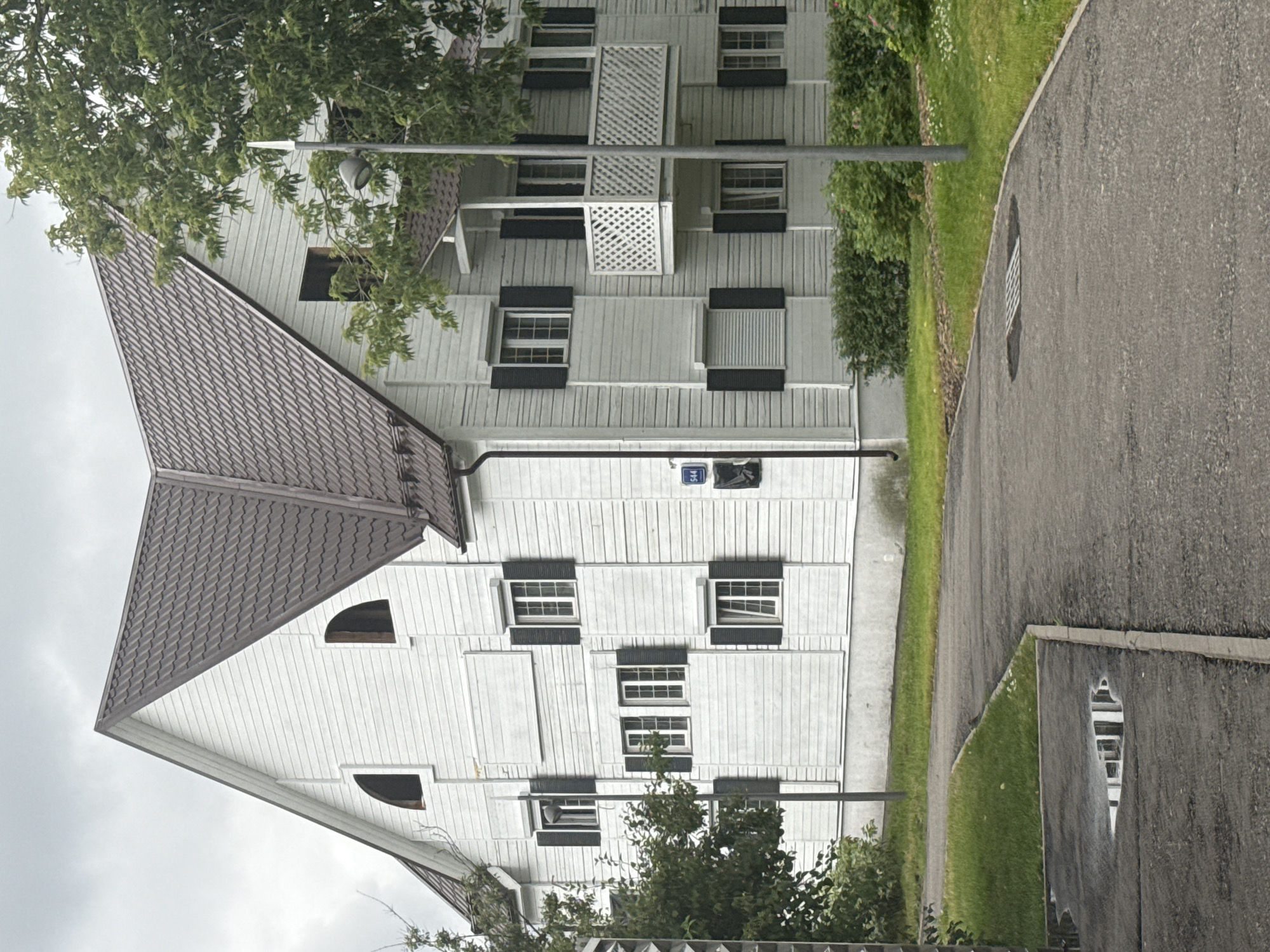 A white, multi-story wooden house with a dark, pitched roof is situated in an outdoor setting under a cloudy sky. The house features numerous windows with dark shutters, a small balcony with a white lattice railing, and is surrounded by greenery, including trees and bushes. A grey asphalt path leads towards the house, with a grassy area adjacent to it. A street light stands near the house. A small puddle reflecting the building is visible on the path. The scene appears to be a residential area within Moscow, Russia, likely a suburban or dacha-style property, given the architectural style and surrounding vegetation. No people are visible in the image.