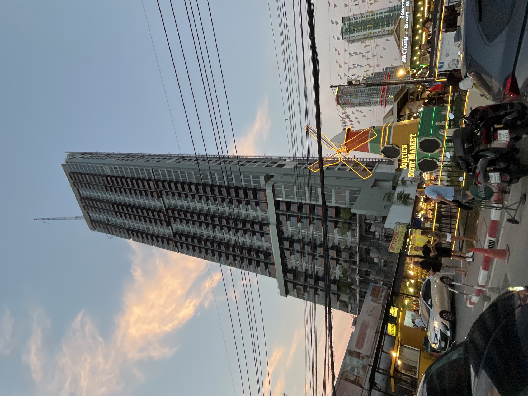 The photograph shows a bustling street scene at dusk in Pattaya City, Thailand. In the foreground are power lines crisscrossing the sky in front of a tall building with a repeating pattern of windows and balconies. To the right, an area with a large windmill lit up with orange lights marks the "Jomtien Night Market," with various stalls and shops with bright lights. People are walking along the sidewalk in front of the market. Cars and scooters are parked or moving in the street. A second, taller building rises in the background, decorated with colorful pennants.
