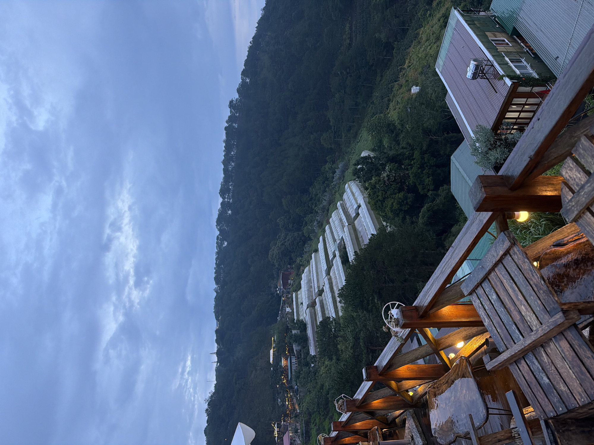 The image captures an elevated view of a hilly landscape under a cloudy, overcast sky. In the foreground, a wooden deck with rustic tables and chairs is visible, some of which appear wet. Several warm-toned lights are strung along the railing, adding a soft glow to the scene.
Beyond the deck, a series of white-tarped structures, likely greenhouses, are arranged in rows on a hillside. These are surrounded by dense green vegetation and trees. Further in the distance, the landscape continues to be covered by a thick forest that stretches to the horizon. Small buildings with illuminated windows and lights are scattered throughout the hillside, suggesting a village or resort. On the right side of the image, a green-roofed building with multiple windows and a water tank on its roof is prominent.
The overall atmosphere is serene and slightly melancholic, possibly due to the cloudy weather and twilight setting. The location appears to be a scenic viewpoint overlooking a cultivated valley and forested mountains. The presence of greenhouses suggests agricultural activity in the area.
