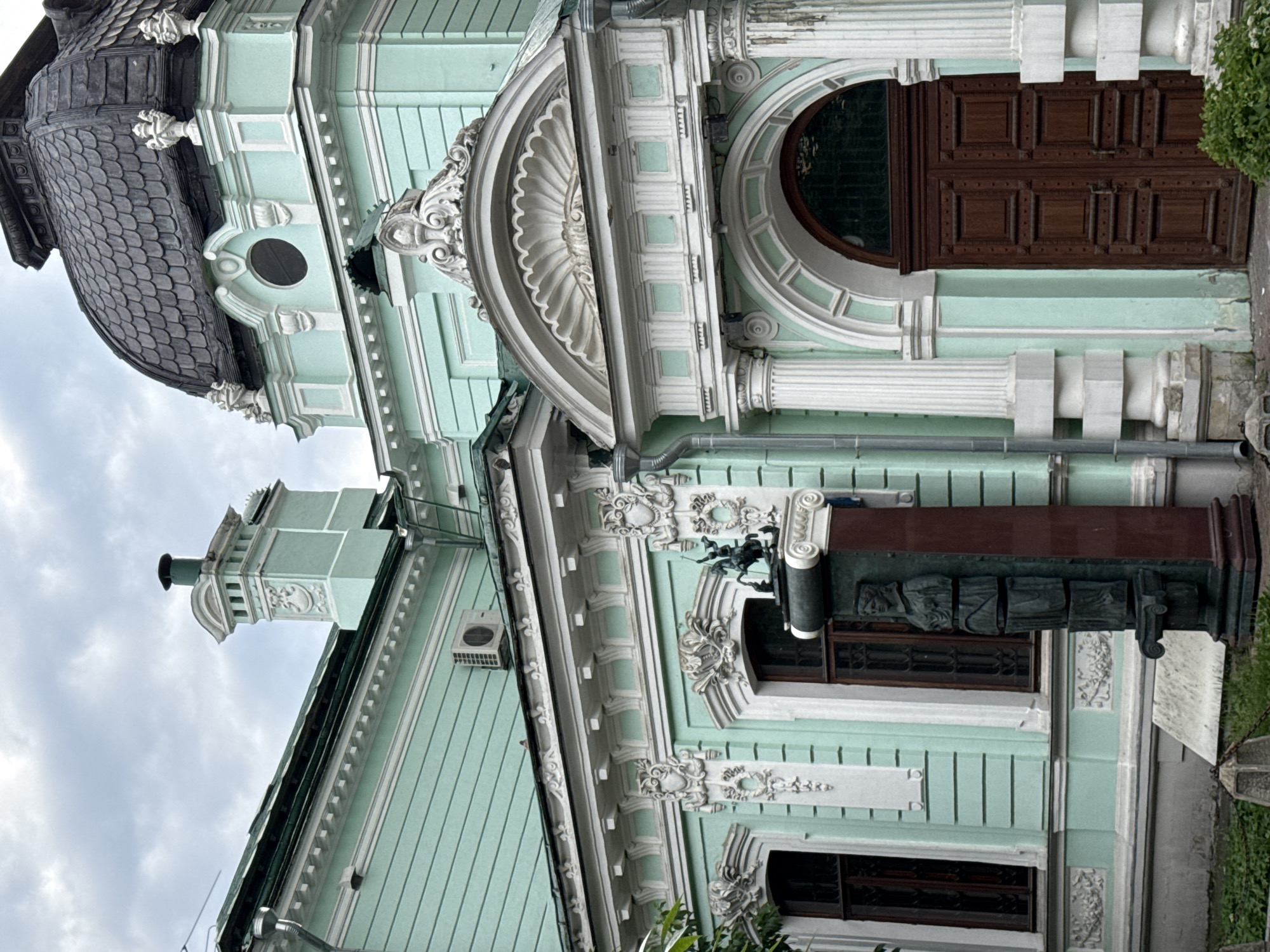 The image captures a detailed view of the exterior of a building with distinct architectural elements. The building is painted in a pale green color and features ornate white trim and decorative carvings. Prominent features include large wooden doors with a grid of square panels, arched windows with rounded tops, and classical columns. The roof is covered in dark, overlapping shingles, and a dome-like structure is visible. Decorative gargoyle-like figures and intricate friezes adorn the facade. A section of the building has a more modern appearance with an air conditioning unit mounted on the wall.
One section of the building includes a monument with a dark red base and a series of bronze sculptures or reliefs depicting figures in draped clothing. The sky above is overcast with clouds, suggesting a possibly cool or temperate day. The image is oriented vertically, making it appear as if the building is leaning.
While no people are visible in the frame, the architecture itself suggests a historical or culturally significant location, possibly a museum, government building, or a grand residence in Moscow, Russia. The overall impression is one of historical grandeur and intricate craftsmanship. No visible text is present in the image.