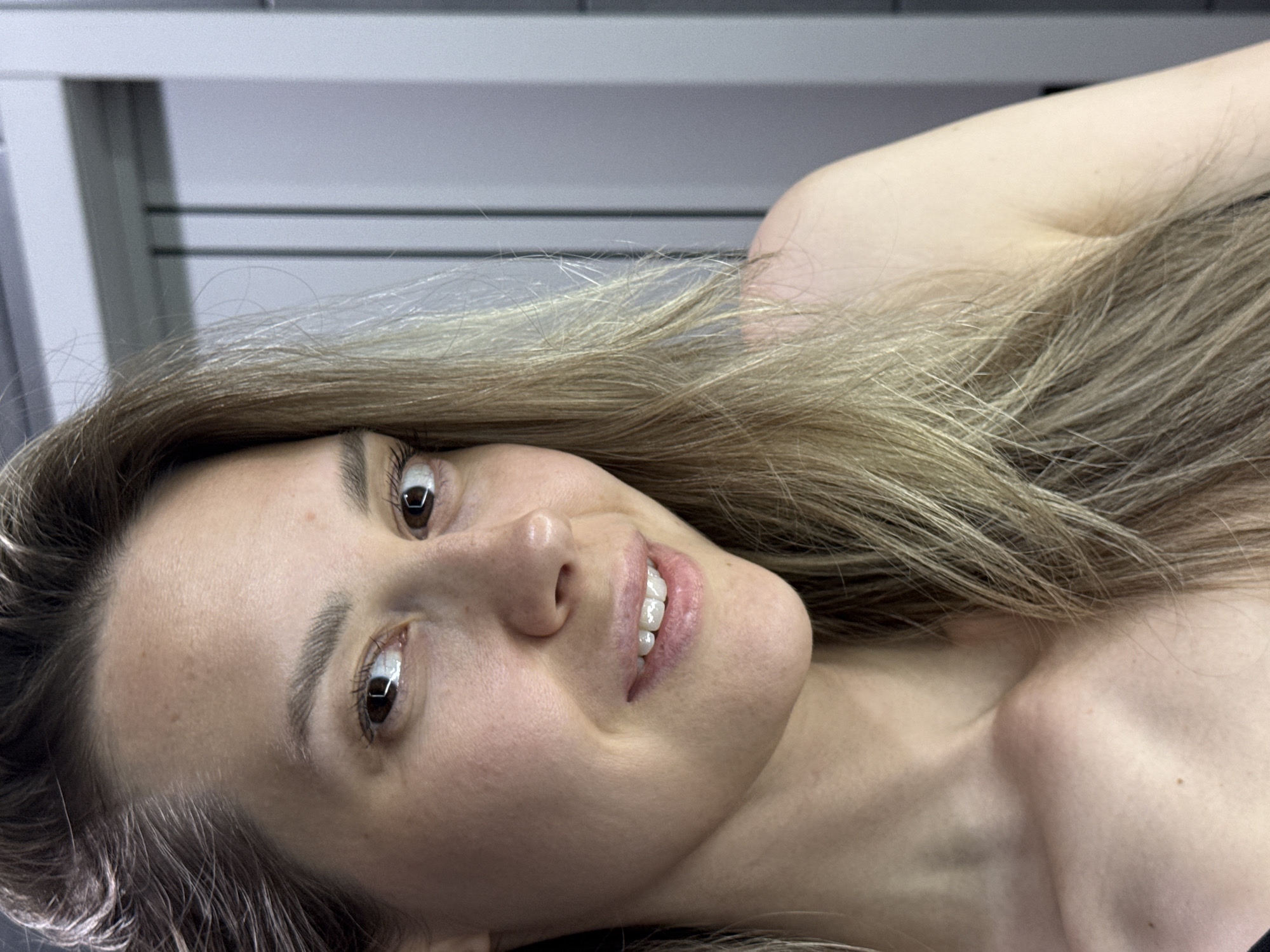 A medium shot depicts a woman with light brown hair, light skin, and brown eyes looking at the camera with a slight smile. The setting appears to be indoors, evidenced by a wall with horizontal lines in the upper left background. The woman's neck and shoulder are visible, suggesting the photo is either a close-up portrait or a selfie. No specific details indicate the location is Chaikovsky, Russia.