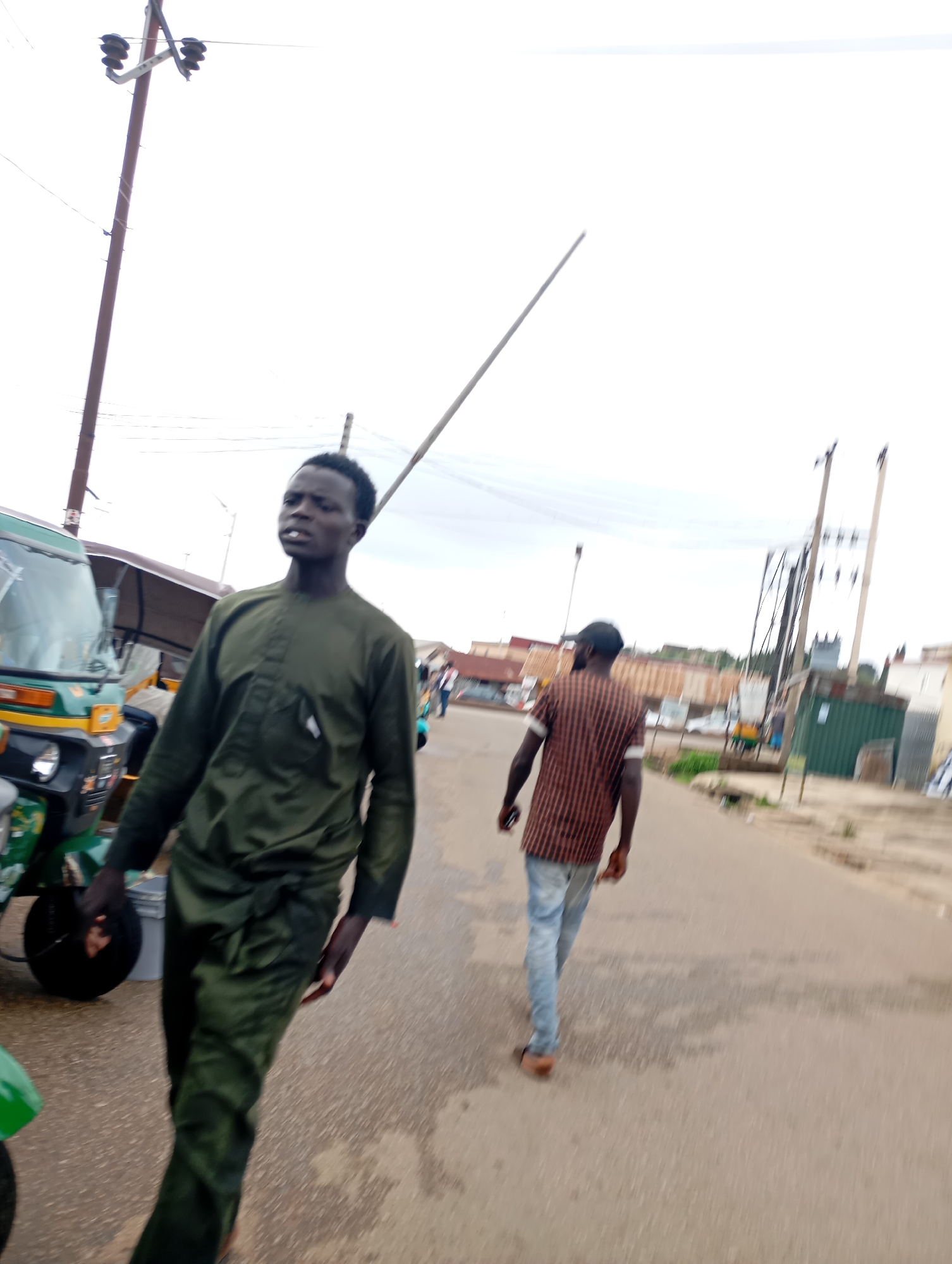 In Jos, Nigeria, a man in a dark green traditional outfit walks towards the camera on a paved road. His left hand is extended, possibly holding a small grey object. Beside him, to his left, is a green and yellow motorized tricycle. In the background, another man with a striped shirt walks away from the camera, and further back, a few more people and buildings are visible. The sky is overcast, suggesting cloudy weather. Electric poles and wires are present throughout the scene.