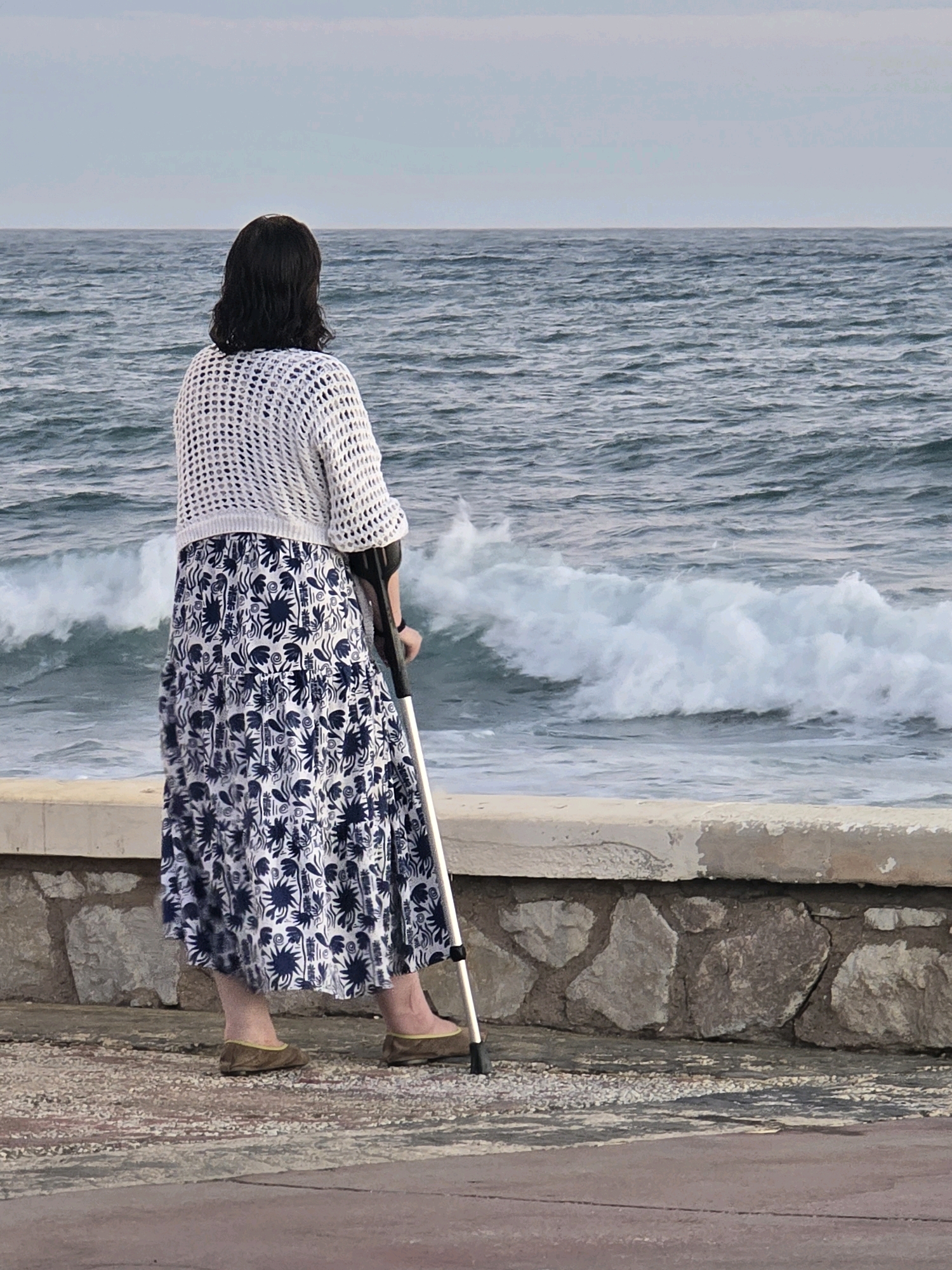 A woman stands on a paved walkway facing a choppy sea. She is wearing a white crocheted sweater and a long, dark blue and white floral print skirt. She is leaning on a black and silver crutch with her right hand. The walkway is bordered by a low stone wall. The sea has white-capped waves. The sky is overcast. The scene appears to be in Málaga, Spain, near the sea.