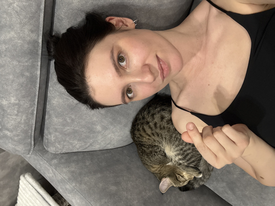 The image presents a close-up, top-down perspective of a person and a cat resting together on a gray fabric couch. The image is oriented such that the person's head is positioned on the left side of the frame, with their body extending towards the right.
The person, likely female, has dark hair pulled back, revealing a clear forehead. They have light skin and prominent, warm brown eyes that are looking directly into the camera. A soft, subtle smile plays on their lips. They are wearing a simple black spaghetti-strap top or camisole. A small, silver hoop earring is visible in their left ear. Their left hand is visible near their chest, performing a 'finger heart' gesture, where the index finger and thumb touch to form a small heart shape.
Cuddled closely next to the person's chest and shoulder area is a small, striped tabby cat. The cat is curled up tightly in a sleeping position, with its head tucked in and its ears slightly visible. Its fur displays a characteristic pattern of dark brown and tan stripes and spots.
Both the person and the cat are situated on a plush, light gray fabric couch. The texture of the upholstery is clearly visible, showing a slightly worn or mottled pattern. The background beyond the couch is indistinct and out of focus, suggesting an indoor setting.
The overall scene conveys a feeling of peace, comfort, and affection. The person appears relaxed and content, sharing a quiet moment with their sleeping pet, with the finger heart gesture adding a tender detail.