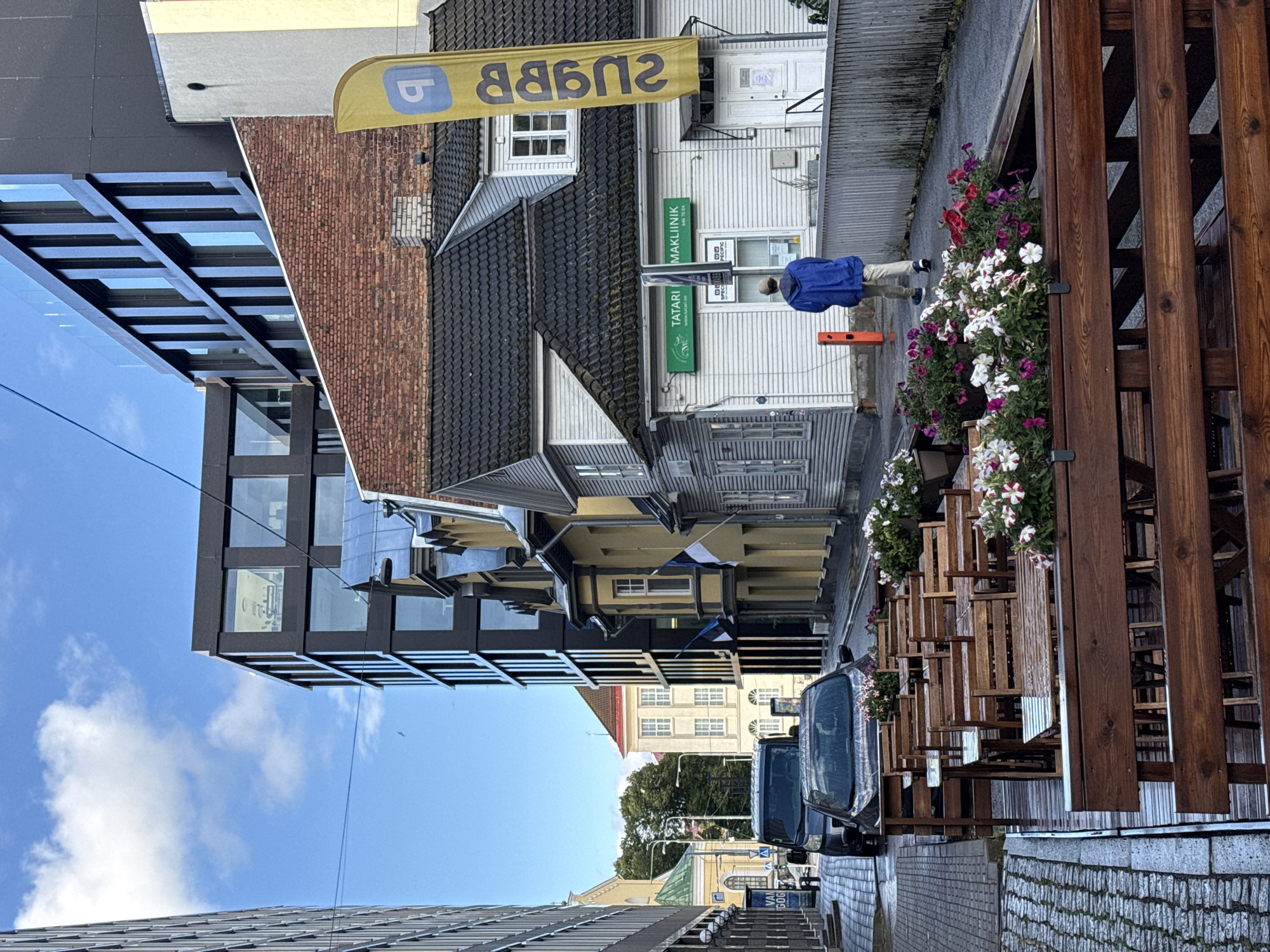 The image depicts a street scene in Tallinn, Estonia. In the foreground, there is an outdoor seating area for a restaurant, with wooden tables and chairs. The tables and chairs are wet, suggesting recent rain. Beyond the seating area, there are several buildings of varying architectural styles. On the right, a white, single-story building has a green sign that reads "TATARI MAAKLIINIK" and a yellow flag with a blue symbol and text. A person wearing a blue hooded jacket and light-colored pants is walking away from the camera towards this building. In the middle ground, a black van and a dark-colored car are parked on the street. Behind these vehicles, several older buildings with colorful facades and Estonian flags are visible. In the background, modern, multi-story buildings with dark facades and large windows dominate the skyline. The sky is blue with scattered clouds. The street is paved with cobblestones.