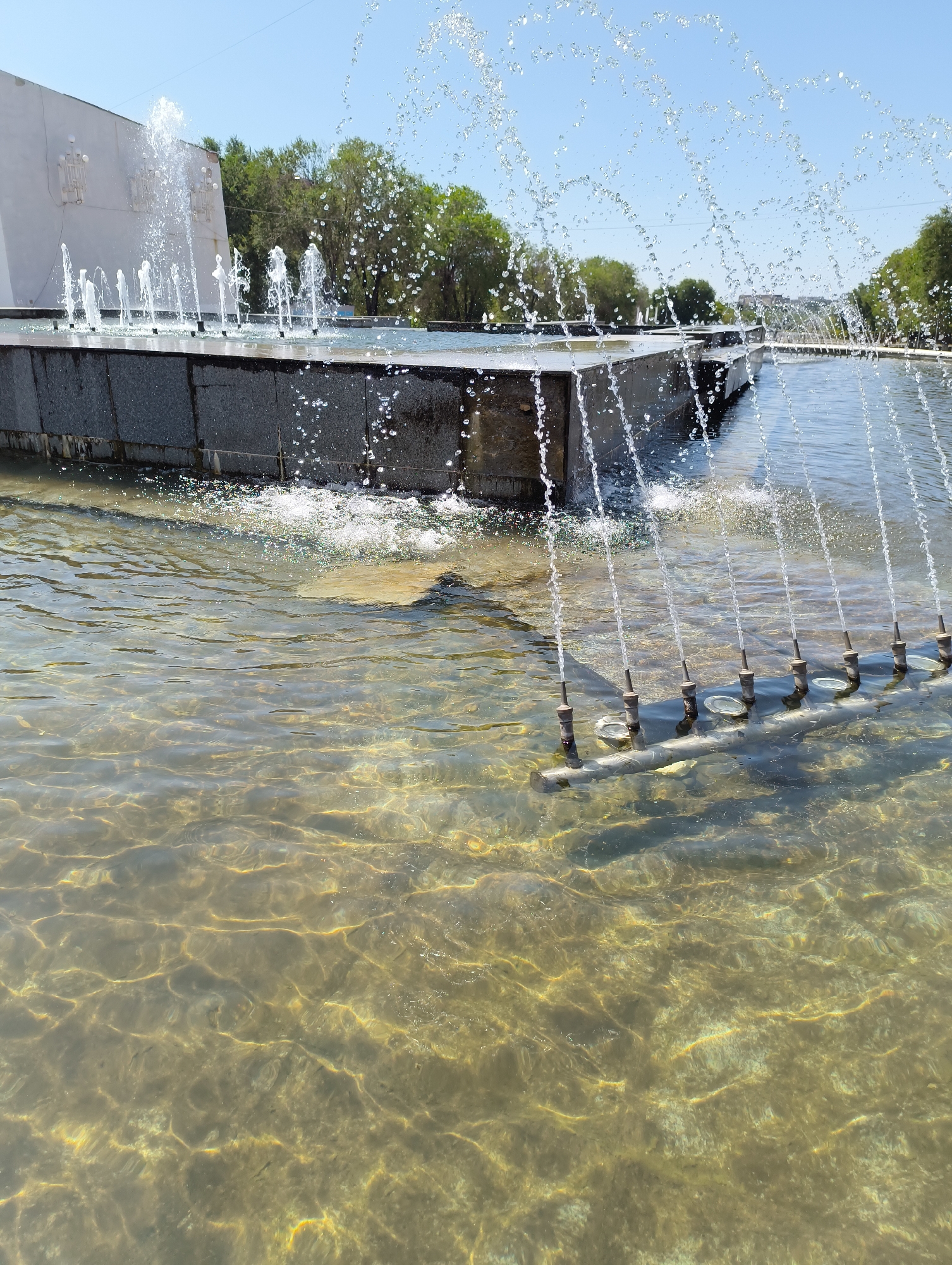 A low-angle, eye-level shot captures a public fountain on a bright, sunny day. The foreground is dominated by a shallow body of water with visible ripples and light reflecting off the surface, creating patterns of golden yellow and brown. Submerged in the water, a metal pipe with multiple nozzles is visible, each directing a thin stream of water upwards.
In the mid-ground, the fountain's tiered structure is evident, with dark, textured stone forming its edges. A line of smaller, vertical water jets emerges from this structure, creating a graceful arch of water droplets against the clear blue sky. Further back, a larger, more powerful jet of water arcs significantly higher.
The background features a white, monolithic building with decorative sconces on its facade. Behind the building, a dense line of green trees forms a natural backdrop, with the leaves illuminated by the sun. The overall scene conveys a sense of calm and recreation, typical of a public park or plaza. The location is identified as Unknown, Unknown.