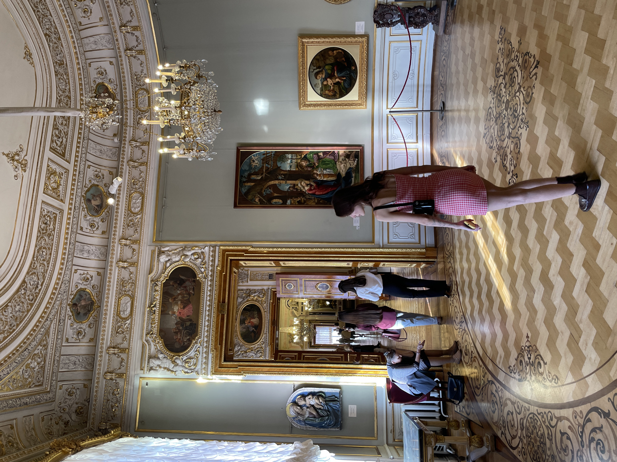 The image shows a room in what appears to be the Winter Palace of the Hermitage Museum in Saint Petersburg, Russia. The room is ornately decorated with a crystal chandelier, paintings, and an intricately designed ceiling with gold detailing. The floor is wooden with a decorative pattern. There are several people in the room: one woman stands near a painting wearing a red and white checkered dress and two other people are standing together, looking at artwork. The light is bright and seems to be coming through the window. The room is generally well lit, allowing for clear views of the architectural and artistic details.