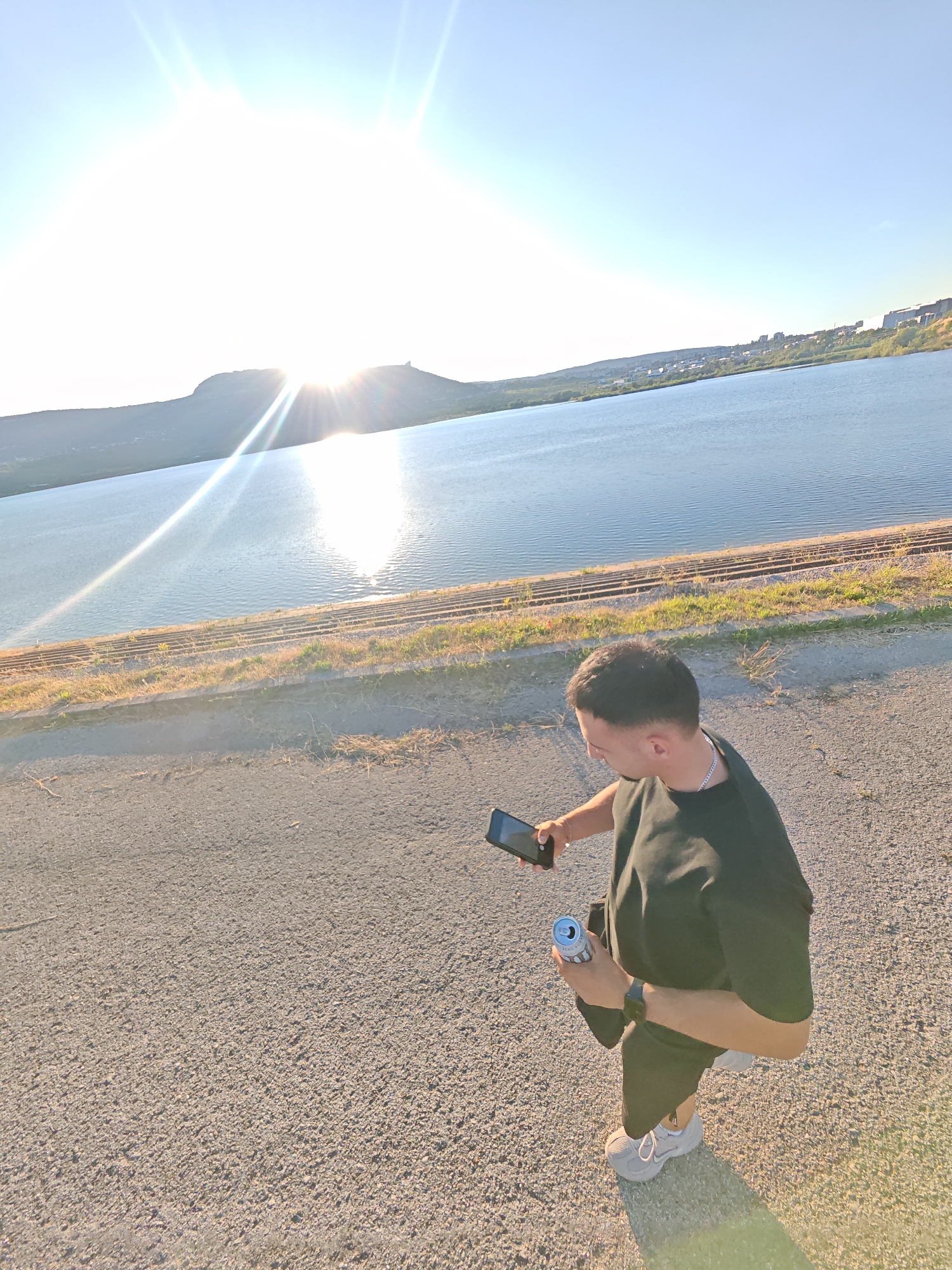 In Shumen, Bulgaria, a young man is captured from a high angle, standing on a paved path beside a large body of water. The sun, a bright orb with strong lens flares, shines from the left, illuminating a distant hill and a portion of the water. The man, wearing a black t-shirt and shorts, holds a smartphone in his left hand and a silver can in his right. He appears to be looking at his phone. The water is calm, reflecting the sunlight. Railway tracks run along the edge of the water, bordered by dry grass and some sparse vegetation. In the background, a town with buildings is visible along the shoreline. The overall scene is bathed in the warm light of a setting or rising sun.