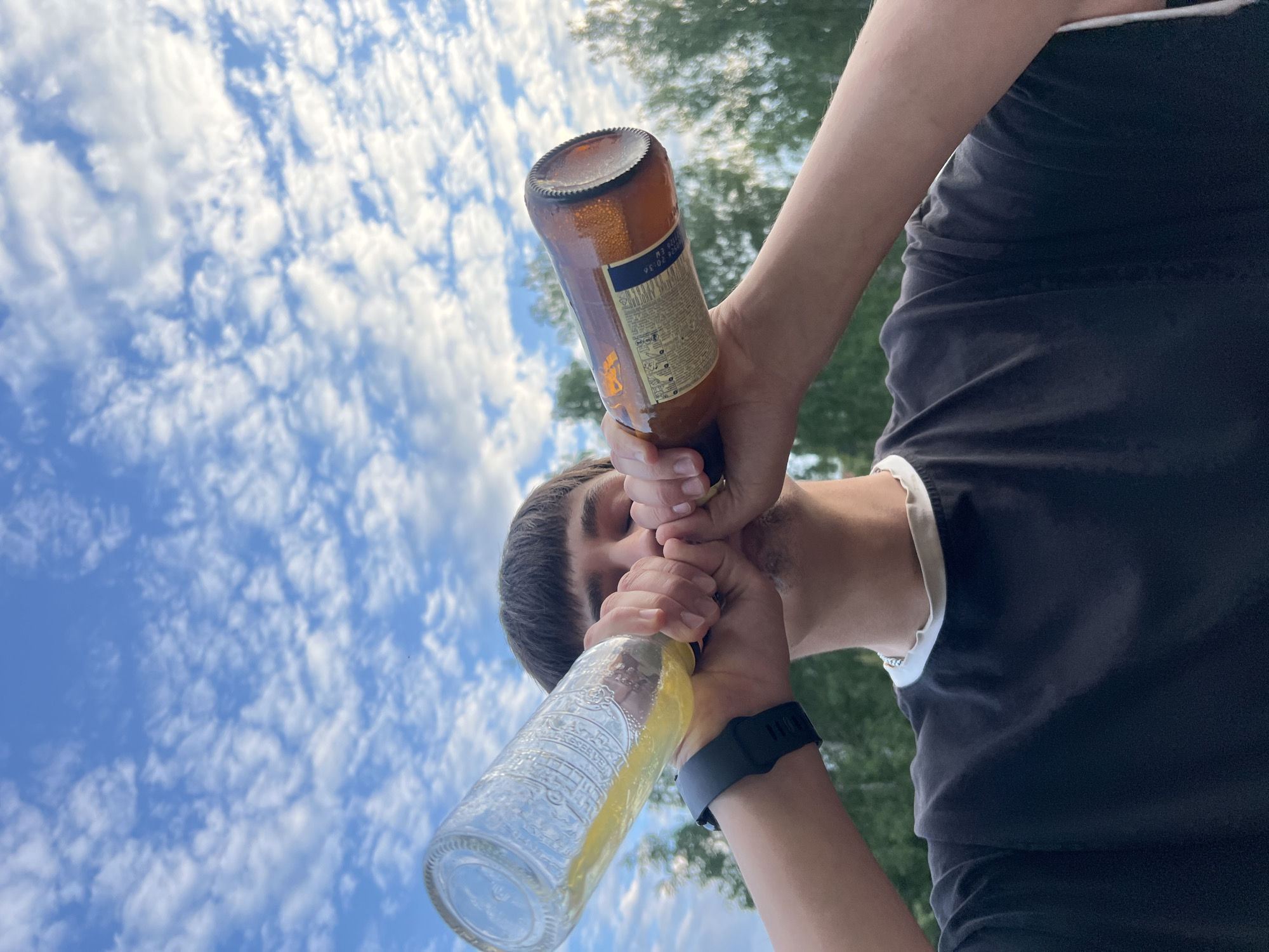 In this low-angle, outdoor shot, a person is captured holding two bottles to their mouth. The sky, filled with wispy white clouds against a bright blue backdrop, suggests a daytime scene. Lush green trees are visible in the blurred background, hinting at a natural or park-like setting. The person appears to be drinking from both bottles simultaneously, with the focus on their interaction with the beverages.
One bottle is made of amber glass and has a label with text and graphics, including what looks like a lion emblem. The other bottle is clear glass and contains a yellow liquid. The person is wearing a black t-shirt with a white trim at the neckline and a black watch on their left wrist. Their face is partially obscured by the bottles, but their eyes are visible and seem to be looking downwards. The overall mood is casual and perhaps playful, given the unusual way the person is holding the bottles. The image is sharpest on the bottles and the person's hands and face, with the background intentionally out of focus. There is no visible text in this description that directly confirms the location as Chaikovsky, Russia, as this information is not present in the visual elements of the image.
