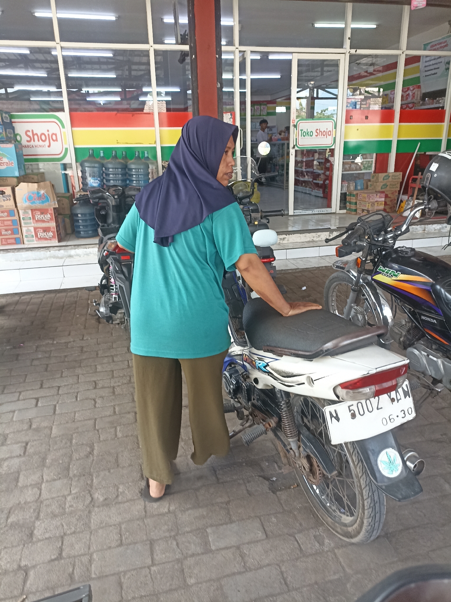 A woman wearing a dark blue hijab and a teal t-shirt over olive green pants stands in front of a storefront in Pandanarum, Indonesia. She is partially obscured by two motorcycles. The storefront, a minimarket, has a large glass facade with brightly colored bands of red, yellow, and green. A sign above the entrance reads "Toko Shoja" with the slogan "HARGA HEMAT" (Economical Price). Inside, shelves stocked with goods are visible. The woman is standing near the handlebars of a white motorcycle, with her right hand resting on its seat. A second motorcycle, black with orange and purple accents, is parked behind and to the right of the first. The ground is paved with grey interlocking bricks. The lighting suggests it is daytime.
