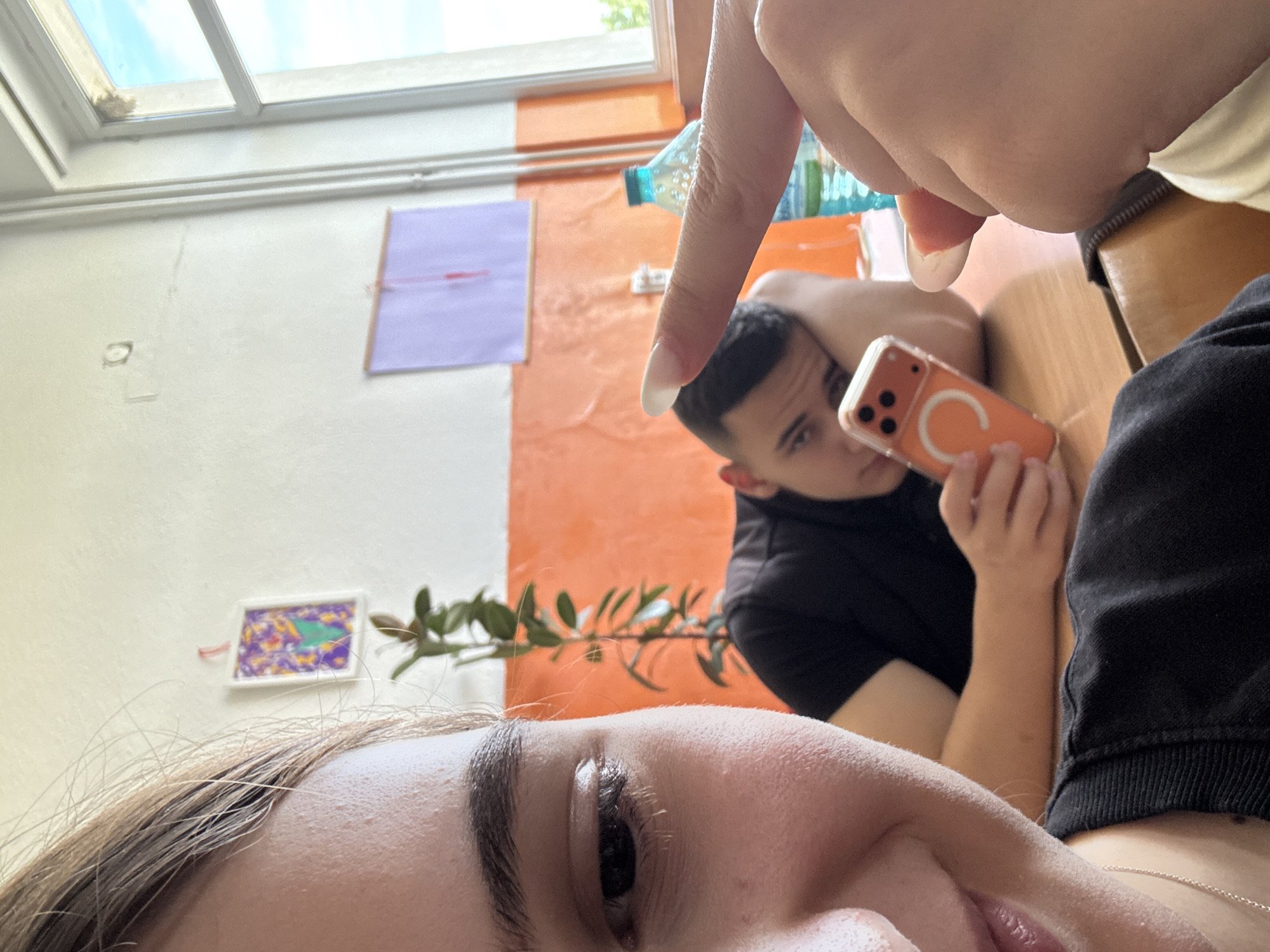 A selfie taken in an indoor environment. A woman's face, including her eye, eyebrow, and hair, is visible in the lower part of the frame. In the background, there is a man holding an orange phone with a MagSafe ring. A finger pointing at the man is visible at the top of the frame. A water bottle is visible behind the finger. The background consists of a wall with a white and orange color scheme, and a window at the top of the frame. A drawing and a purple note are attached to the wall. A plant is also visible in the background. The location is Panagyurishte, Bulgaria.