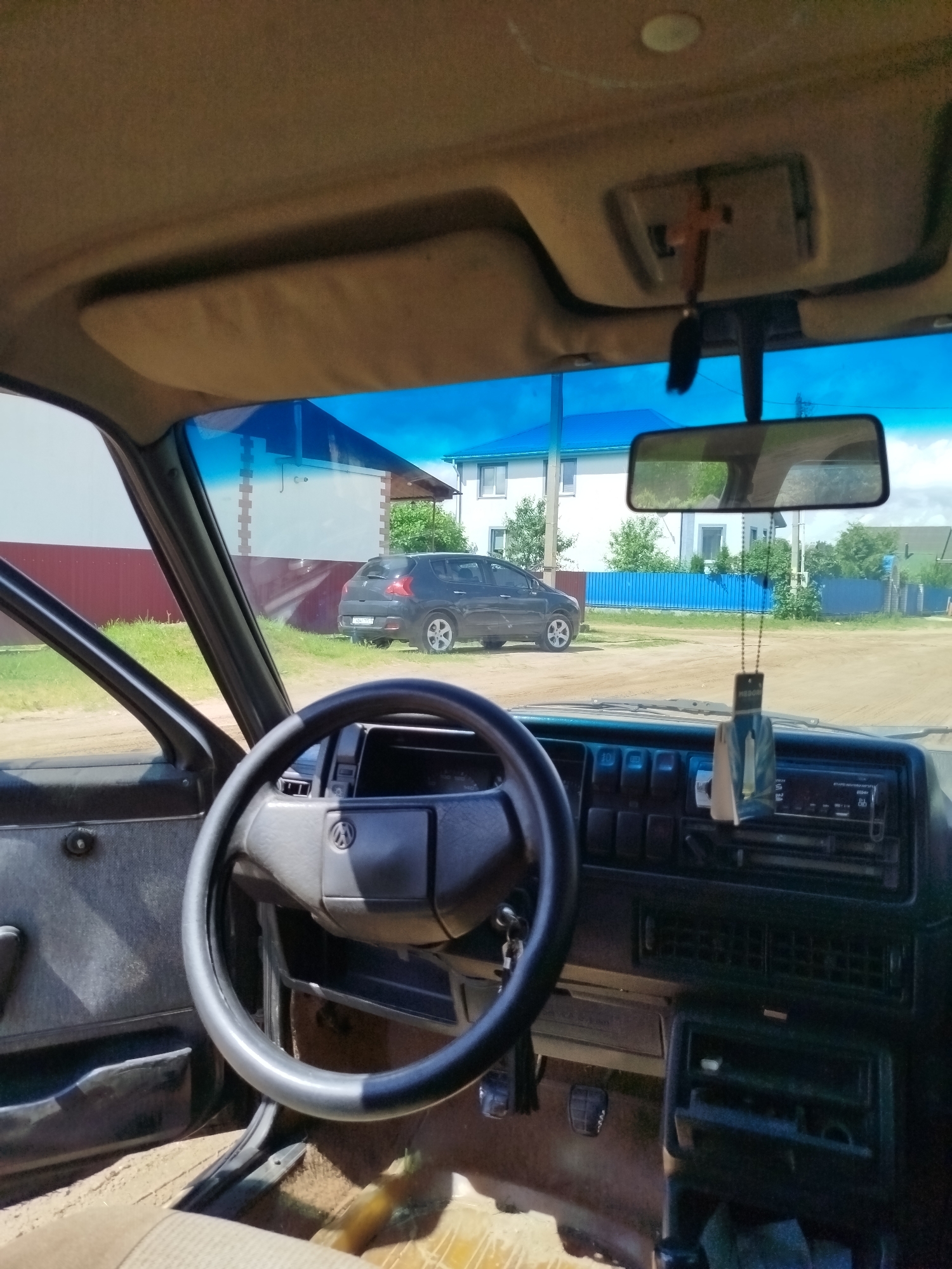 The image shows the interior of a car, looking out through the windshield. The dashboard is visible, including the steering wheel, radio, and air vents. A car air freshener is hanging from the rearview mirror. Outside the car, a dark-colored hatchback is parked on a dirt road in front of a house with a blue fence. The sky is blue with some clouds. The location is identified as Vileyka, Belarus.