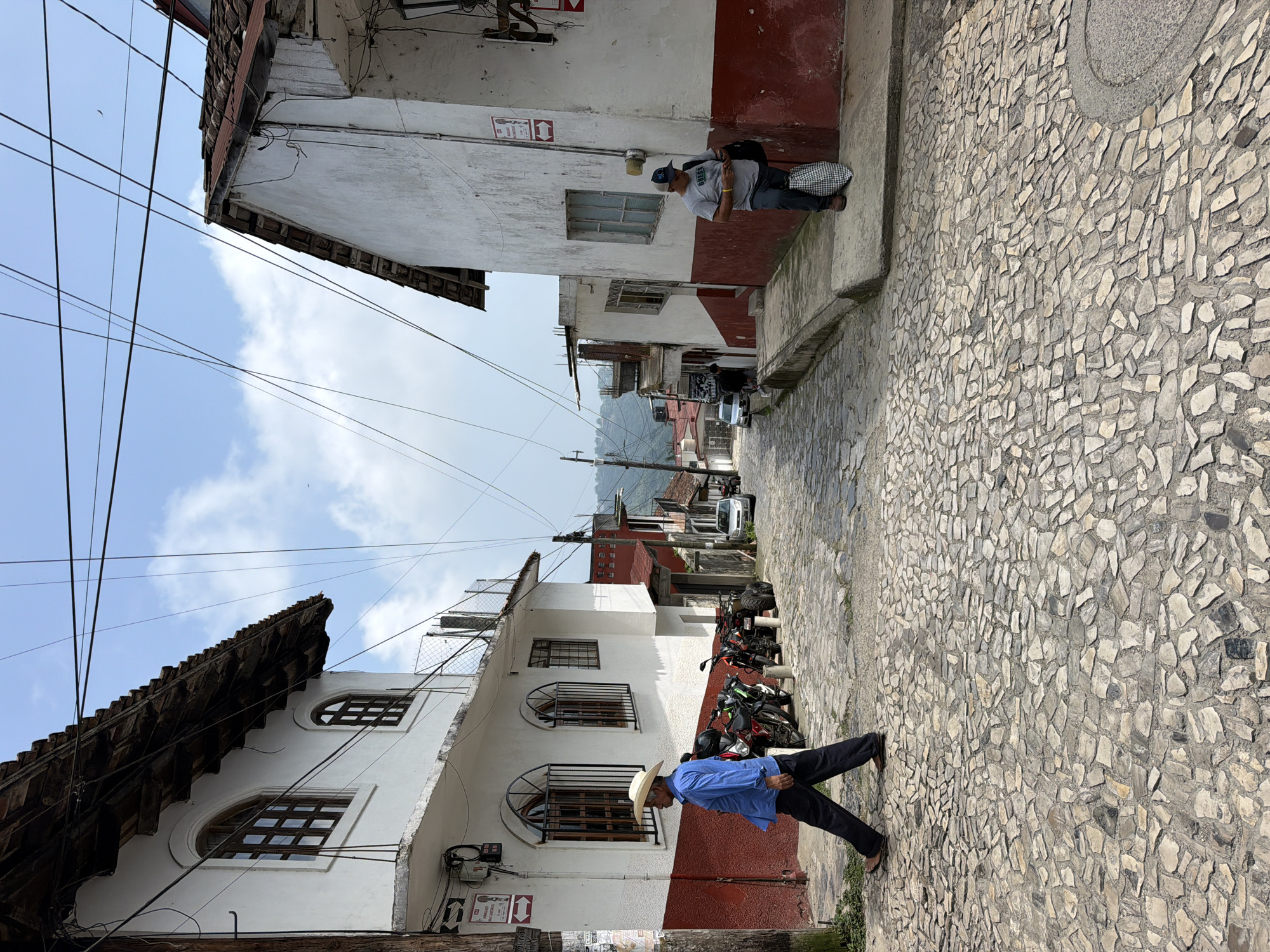 A vertical-format photo shows a street scene in Cuetzalan, Mexico. The street slopes upward away from the viewer, paved with light-colored, irregular stones. The buildings lining the street are two stories, with white walls and red trim on their lower halves. One man wearing a hat and blue shirt is walking on the right side of the street. Another man with a baseball cap, t-shirt, and backpack stands by a building on the left side. Numerous power lines crisscross overhead. Several motorcycles are parked along the left side of the street further up the slope. In the distance, vegetation-covered mountains can be seen. The sky is light blue with scattered white clouds.
