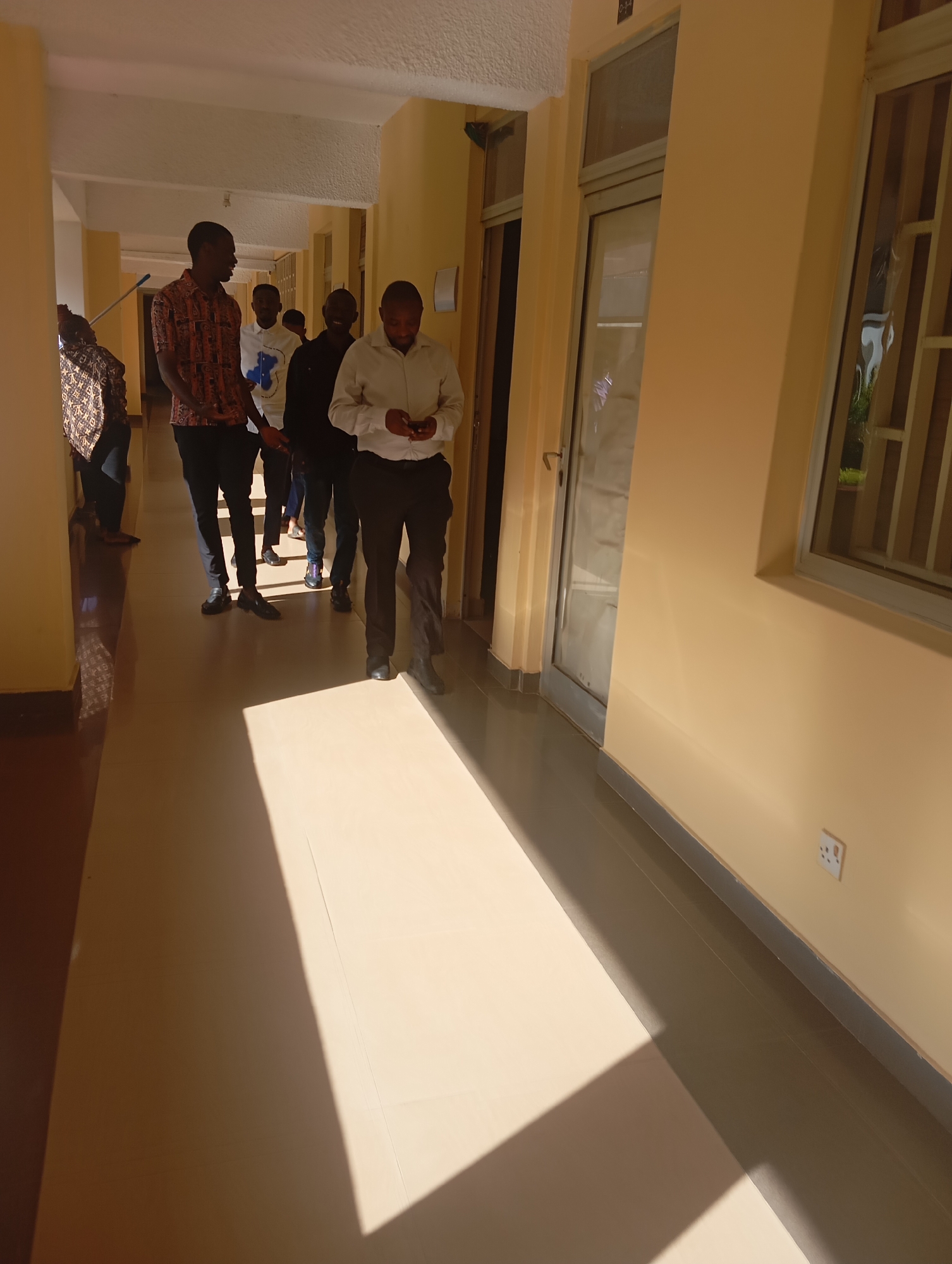 A group of six young Black men and one Black woman are walking down a sunlit hallway in an indoor location. The hallway is wide with cream-colored walls and a light-colored tiled floor. The woman, on the far left, is wearing a patterned top and dark pants and is holding a blue broom. The men are dressed in casual to semi-formal attire, including button-down shirts and pants. They appear to be walking in the same direction, with the first man in the foreground looking down at his phone. The scene suggests a daytime setting, possibly within a school or office building in Jos, Nigeria, given the sunlight casting distinct shadows.
