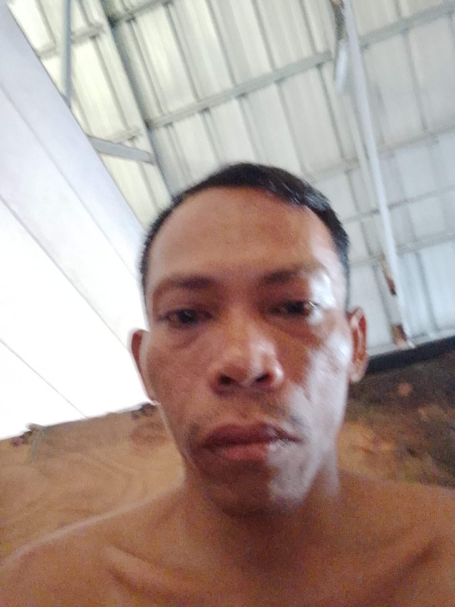 The image is a close-up selfie of an adult male with short, dark hair. He is indoors, underneath a metal and translucent roof structure. The lighting is somewhat diffused. The surrounding area is slightly out of focus. The location is Bukit Ketok, Indonesia.