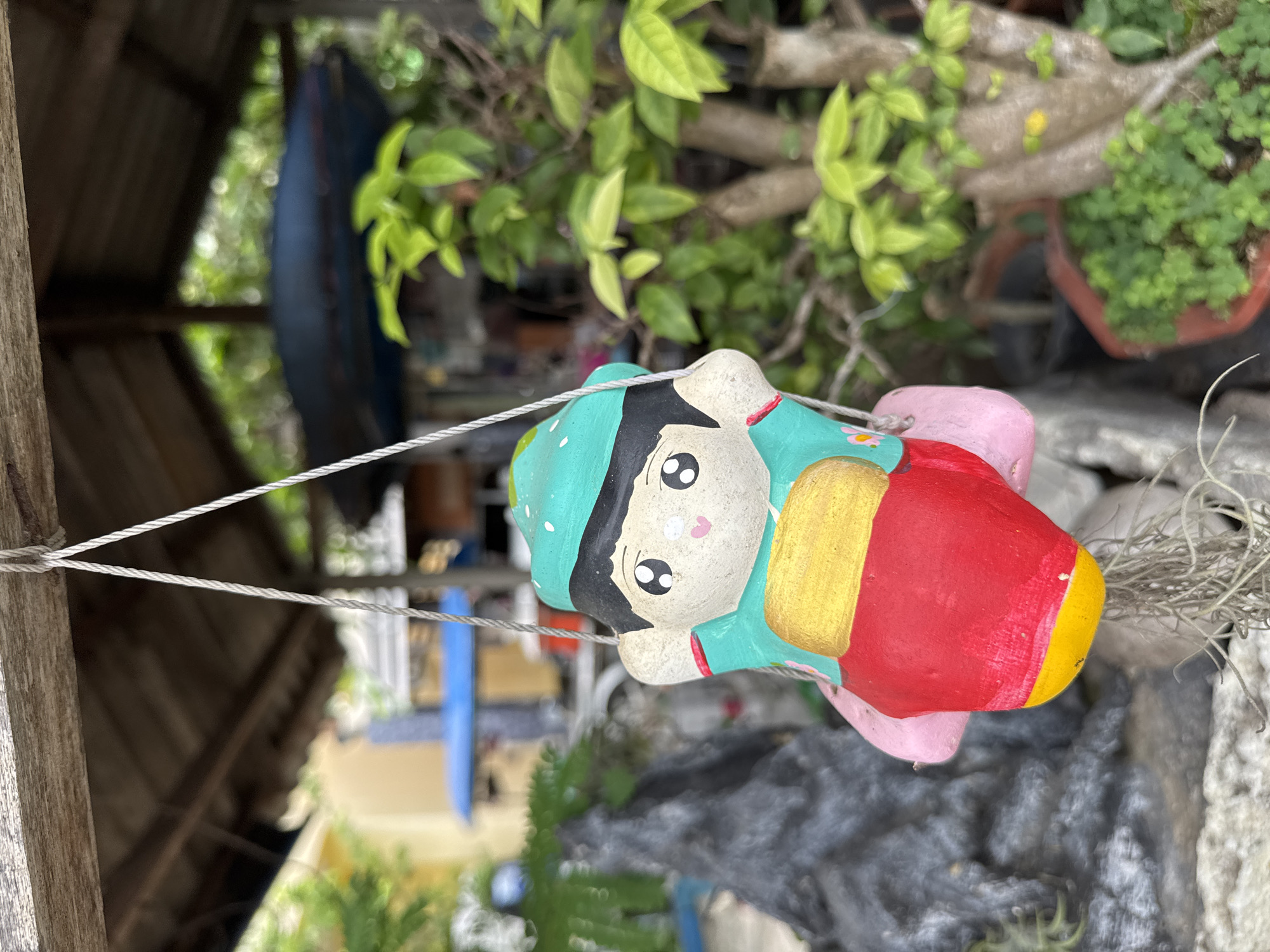 The image shows a small, colorful ceramic figurine of a child hanging from a string. The figurine is wearing a teal hat with white polka dots and a teal outfit with red trim. It has large, wide eyes with black pupils and pink cheeks. The figurine is sitting on a red object, possibly a balloon or a piece of fruit, with yellow and pink accents. The figurine is suspended in front of a blurred background that suggests an outdoor setting. In the background, there are trees with green leaves, a wooden structure that appears to be a roof or awning, and some other blurred objects that could be part of a market or garden. The overall impression is of a whimsical decoration in a casual, outdoor environment. This scene is located in the city of Rawai, Thailand.