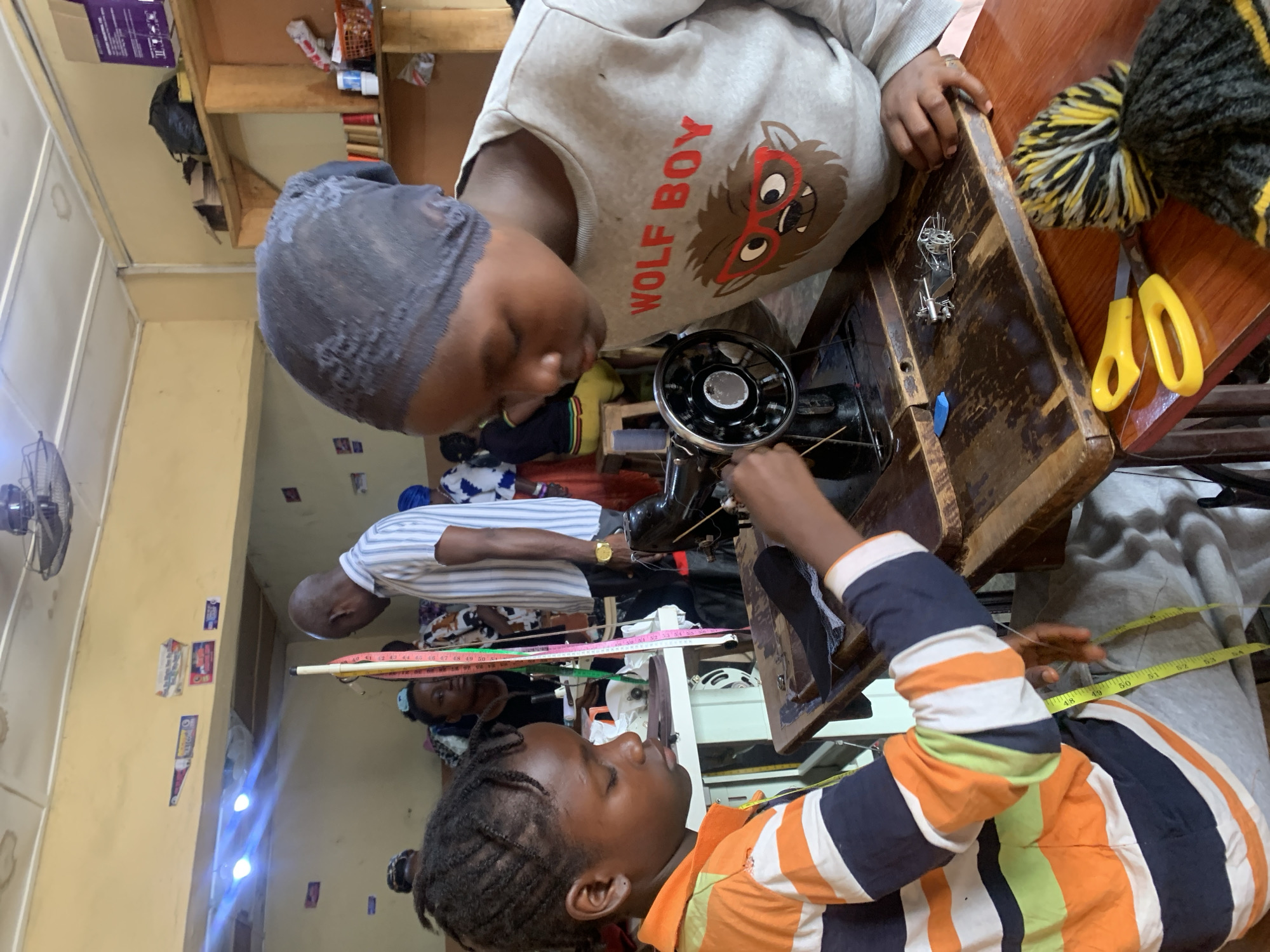 The image shows a group of people in a workshop, likely a tailoring or seamstress shop in Jos, Nigeria. The main subjects are several individuals, some of whom appear to be learning or practicing sewing.
In the foreground, a young person wearing a striped long-sleeved shirt is focused on a sewing machine. Their arms are positioned as if operating the machine, and a measuring tape is visible around their waist, suggesting they are involved in fitting or taking measurements.
To the left and slightly behind, another individual is seated, also appearing to be engaged with sewing. They are wearing a grey sweatshirt with the text "WOLF BOY" and a graphic of a cartoon wolf.
In the background, other individuals are present, one wearing a striped shirt and another in a patterned fabric, further indicating a collaborative or educational environment related to sewing. A sewing machine is prominently placed on a wooden table, along with tools like scissors and a bobbin. The setting appears to be a workshop with shelves in the background holding various items.
The lighting suggests it is daytime, with artificial lights also visible. The overall atmosphere seems focused and industrious, with the individuals concentrating on their tasks. The presence of the measuring tape and sewing machines points to an activity related to garment making or alteration.