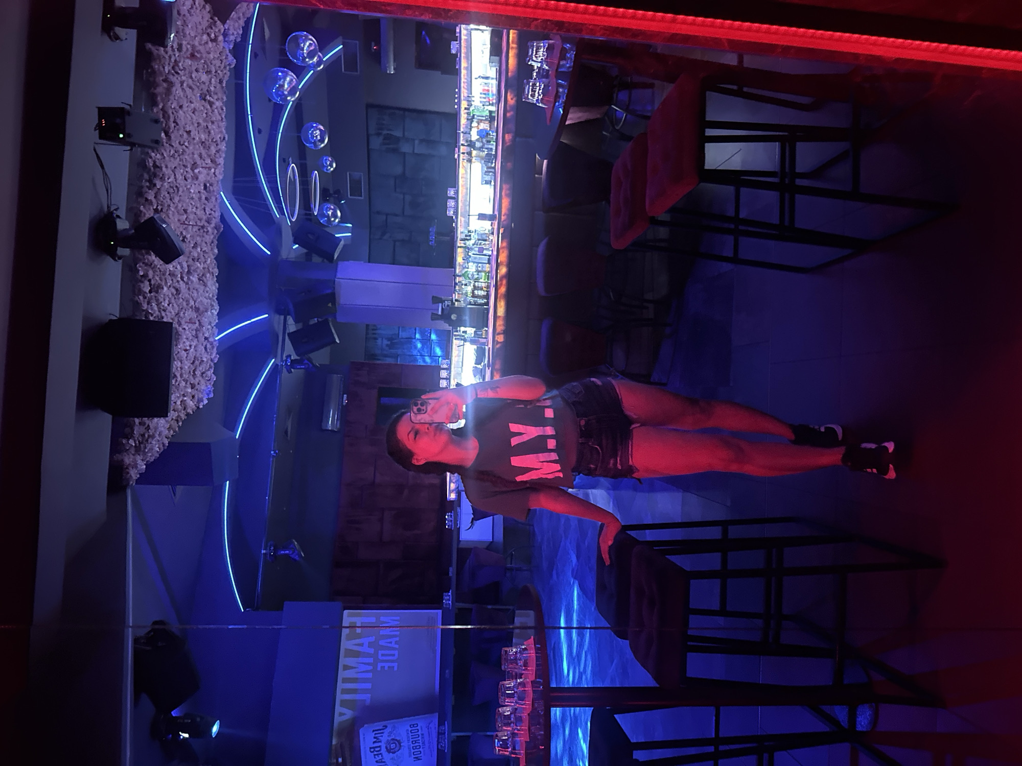 The image depicts a woman taking a mirror selfie in what appears to be a nightclub or bar. The environment is dimly lit, with prominent blue and red neon lighting. The woman is wearing a cropped t-shirt with "M.Y." printed on it and denim shorts. She is standing in front of a mirror, and the reflection shows a bar area with bottles displayed on shelves and stools. In the background, there are disco balls hanging from the ceiling, and spotlights are visible. To the left of the woman, a round table with glasses is visible. The overall ambiance is that of a nightlife venue. The location is identified as a nightclub in Sliven, Bulgaria.