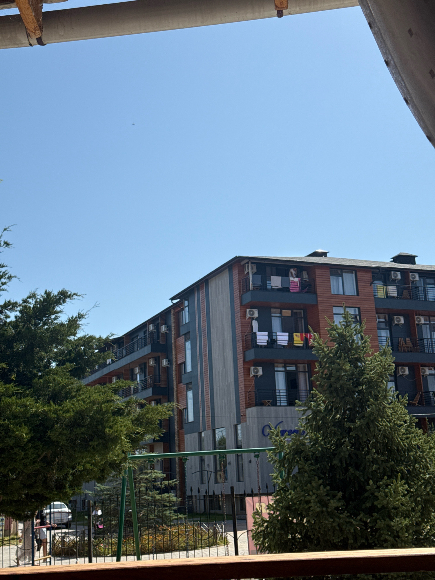 The image shows an apartment building and trees against a clear blue sky. On the balconies of the building, towels are hung out to dry. A swing set is visible in the foreground, along with a fence and some parked cars. A small dark object, possibly a bird or insect, is visible in the sky. The scene is likely a residential area in Bosteri, Kyrgyzstan, given the architecture and vegetation.