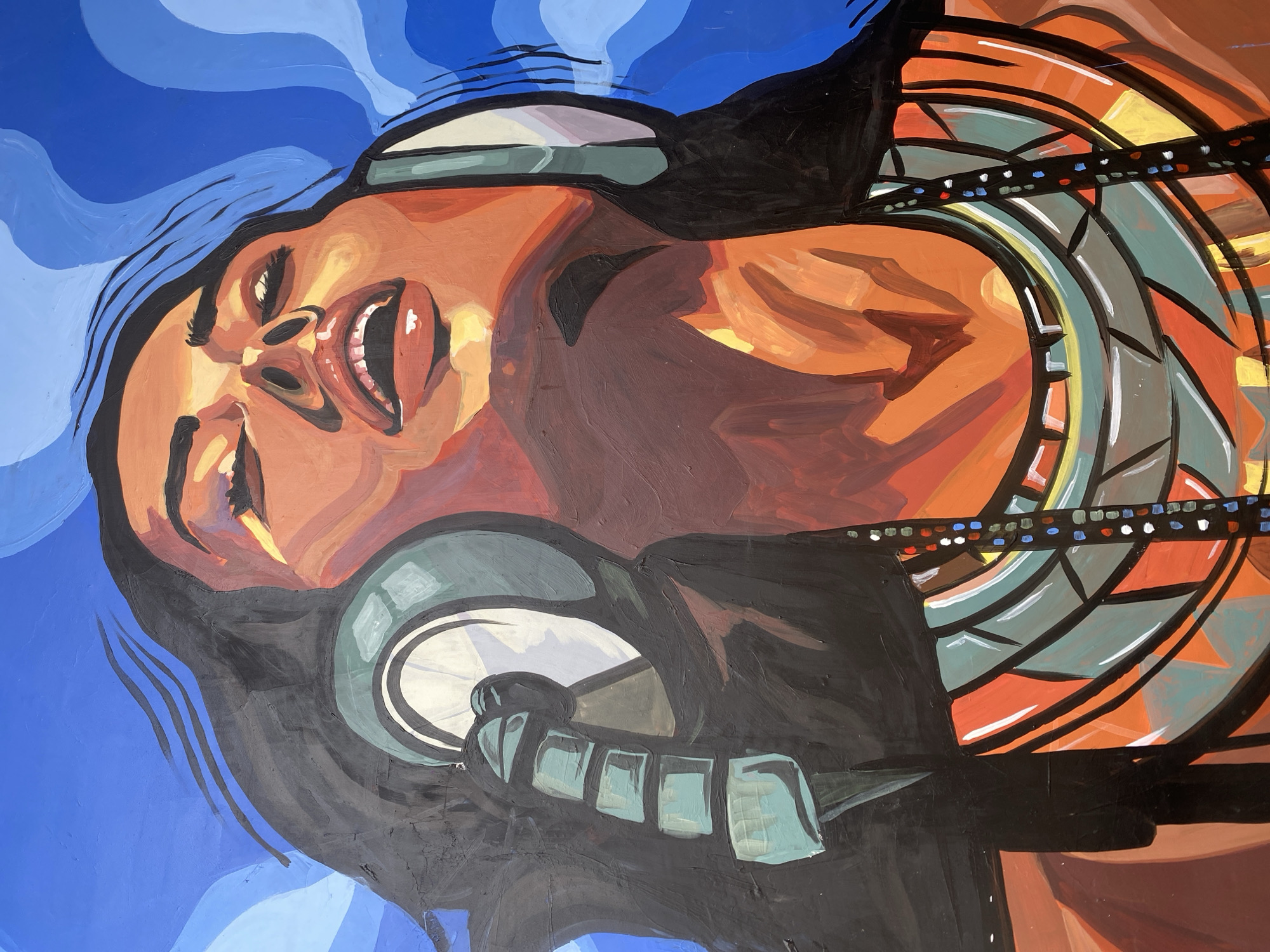 This is a vibrant, stylized painting or mural depicting a woman experiencing intense musical emotion, located in Unknown, Unknown. The woman is shown from the chest up, with her head tilted significantly backward, eyes closed or squinting, and mouth wide open, suggesting she is singing or shouting. Her dark, long hair frames her face and flows upward into the dynamic blue background. She wears large, light blue-green headphones with a visible microphone extending from the left side towards her mouth. Her skin tone is rendered in warm, rich shades of orange, reddish-brown, and yellow highlights, with stark contrasts creating shadows and depth. Around her neck, she wears a striking, multi-layered geometric necklace in shades of teal, orange, gray, and yellow, outlined in black, with an additional strand of small, colorful beads (white, blue, red, yellow) trailing down. The background consists of abstract, wavy shapes in various shades of blue, creating a sense of movement and energy around her head. The artwork features visible brushstrokes and bold, block-like color application.