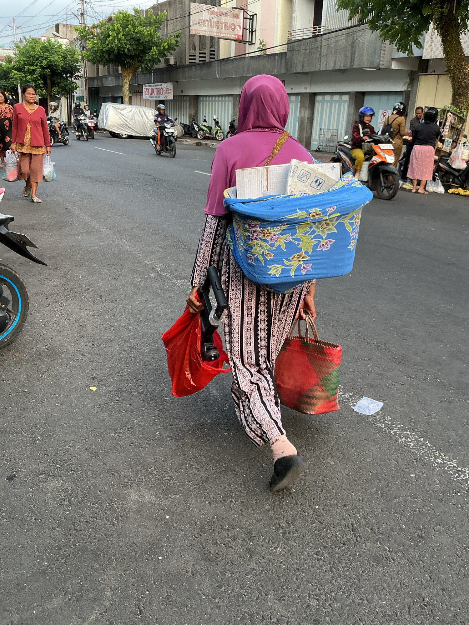 A woman wearing a pink hijab and a patterned dress walks away from the camera on a street in Magelang, Indonesia. She carries a blue woven basket on her back containing items, and in her right hand, she holds a red plastic bag with a black object inside, possibly a vacuum cleaner hose. She also carries a red and green woven bag in her left hand. A motorcycle is parked to her left. In the background, other people on motorcycles and buildings line the street. The scene appears to be a busy commercial or residential area.