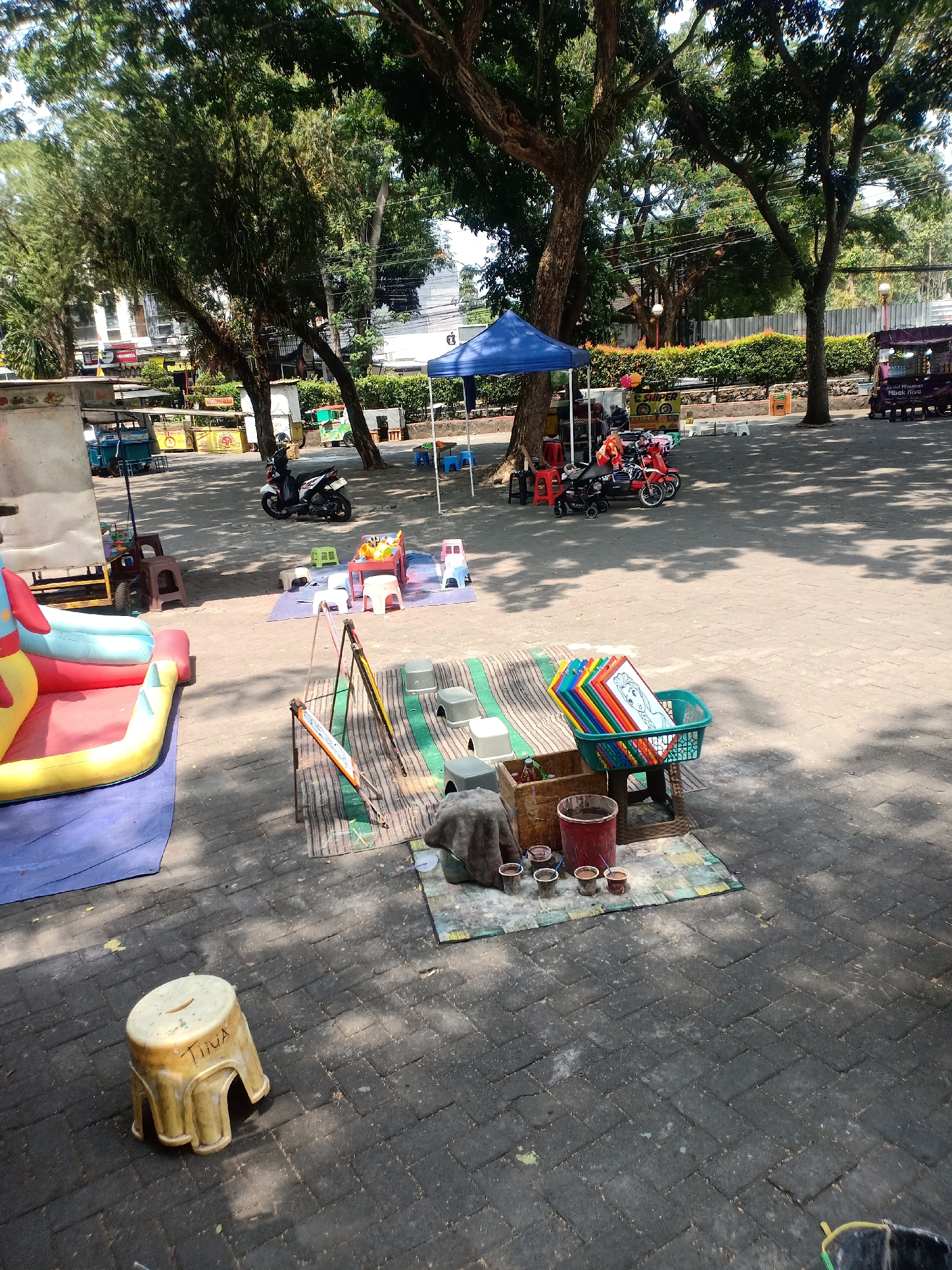 In Semarang, Indonesia, a scene unfolds in a public outdoor area, likely a park or plaza, characterized by a paved ground and ample shade from large trees. A blue tent is set up, suggesting a temporary stall or activity area. To the left, a vibrant, inflatable bouncy castle with red, yellow, and blue sections is visible, indicating a children's play area.
Adjacent to the bouncy castle, a collection of small stools and tables are arranged, some with colorful mats. Further into the scene, a display of children's artwork or coloring pages, featuring characters like Elsa from Frozen, is neatly stacked in a green basket. Below this, small pots containing paint or other art supplies, along with brushes, are laid out on a mat. A red bucket and a wooden crate are also part of this setup.
In the middle ground, several toy motorbikes are parked, some with small children or individuals seated on them, suggesting they are available for rent or play. A white motorcycle is parked nearby. In the background, various food stalls or small shops with colorful awnings are visible, hinting at a lively commercial area. Further back, a fence and lush green bushes line the perimeter, with buildings and trees beyond them. A street lamp is also visible in the distance. The overall impression is that of a recreational space where children can play and families can gather, possibly during a market day or a local event.