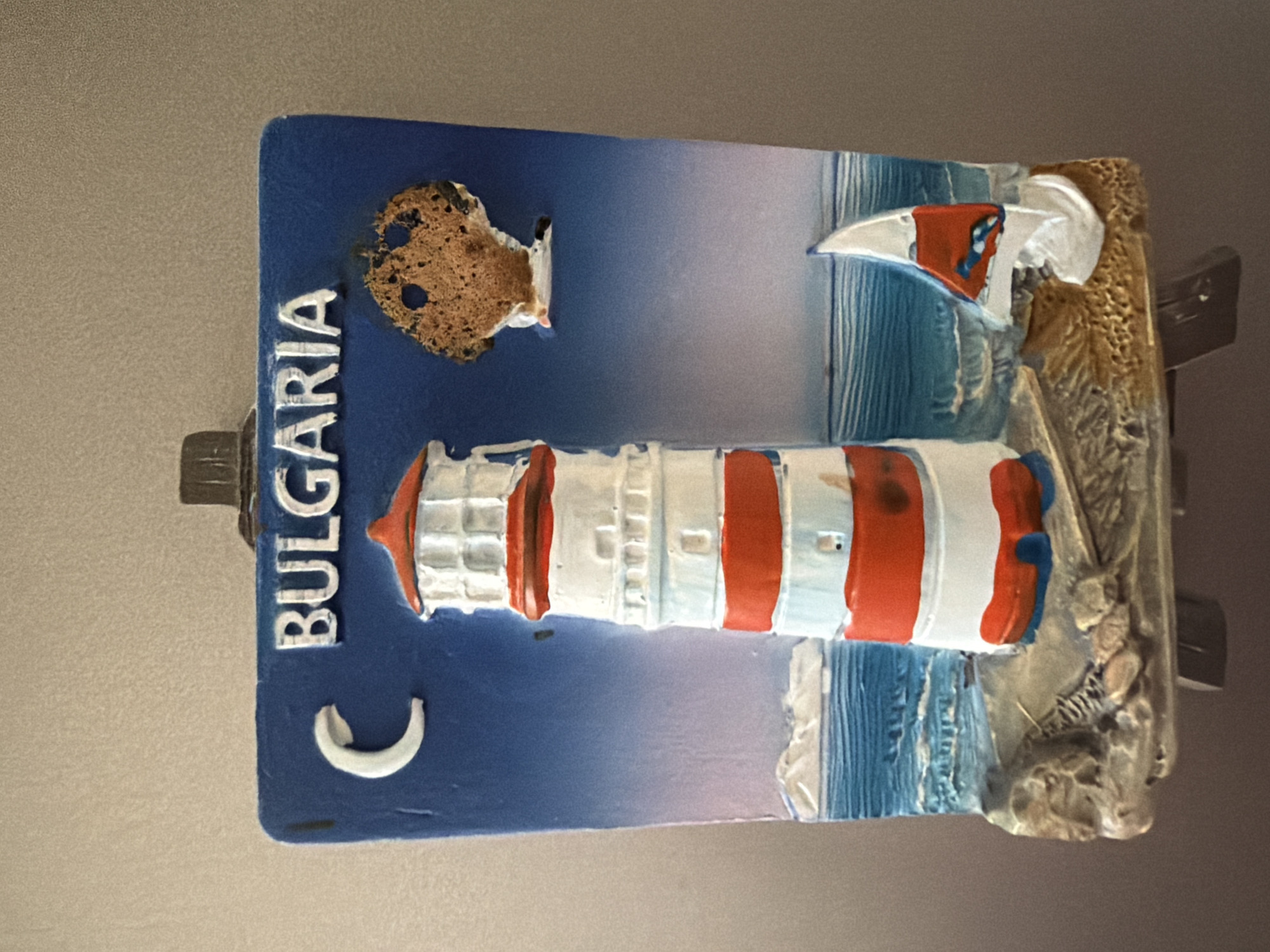The image displays a souvenir magnet shaped like a lighthouse on a rocky shore with a sailboat. The background depicts a gradient sky from blue to pink, with a crescent moon on the left. The word "BULGARIA" is written in white, raised lettering across the top of the magnet. The lighthouse is red and white striped, and the sailboat is white with a red and blue sail. The magnet appears to be attached to a larger display or stand. There is no indication of the city of Pazardzhik in the image.