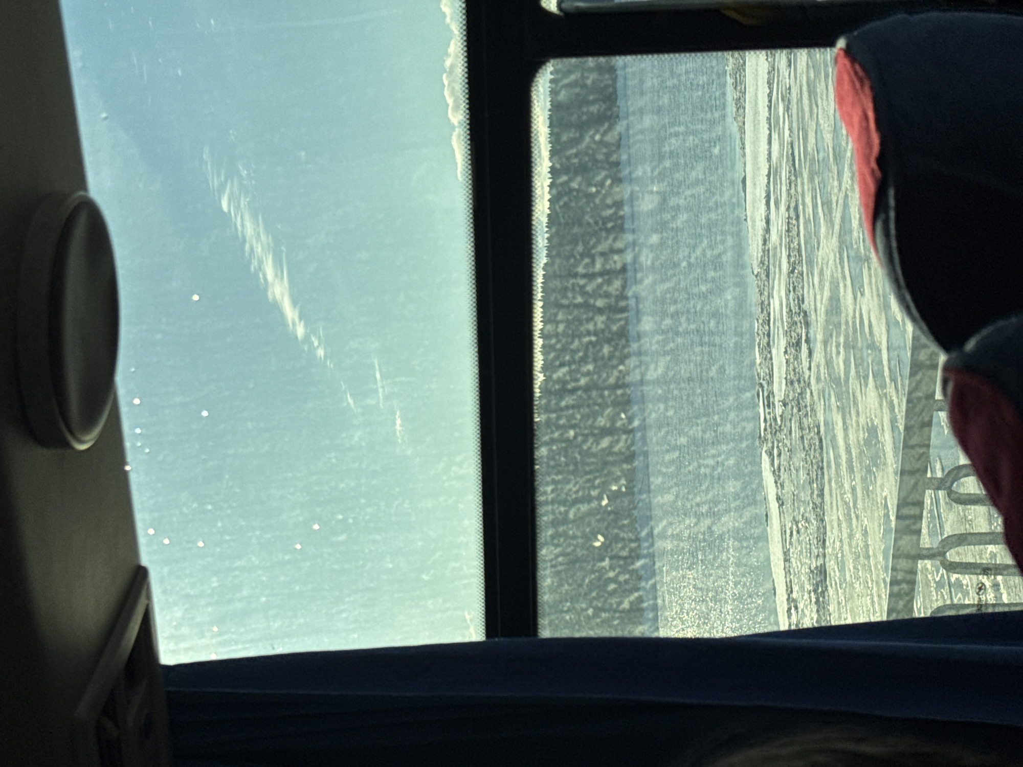 The image shows a view from inside a vehicle, likely a bus or train, looking out of a window. Outside, there is a winter scene. The landscape appears to be covered in snow and ice. The window on the right is partially covered with frost or condensation, obscuring the view. In the background, there are trees covered in snow. A headrest from the vehicle is visible on the right, with a red and black color. The location is likely Usolye, Russia, but this cannot be definitively confirmed from the image alone.