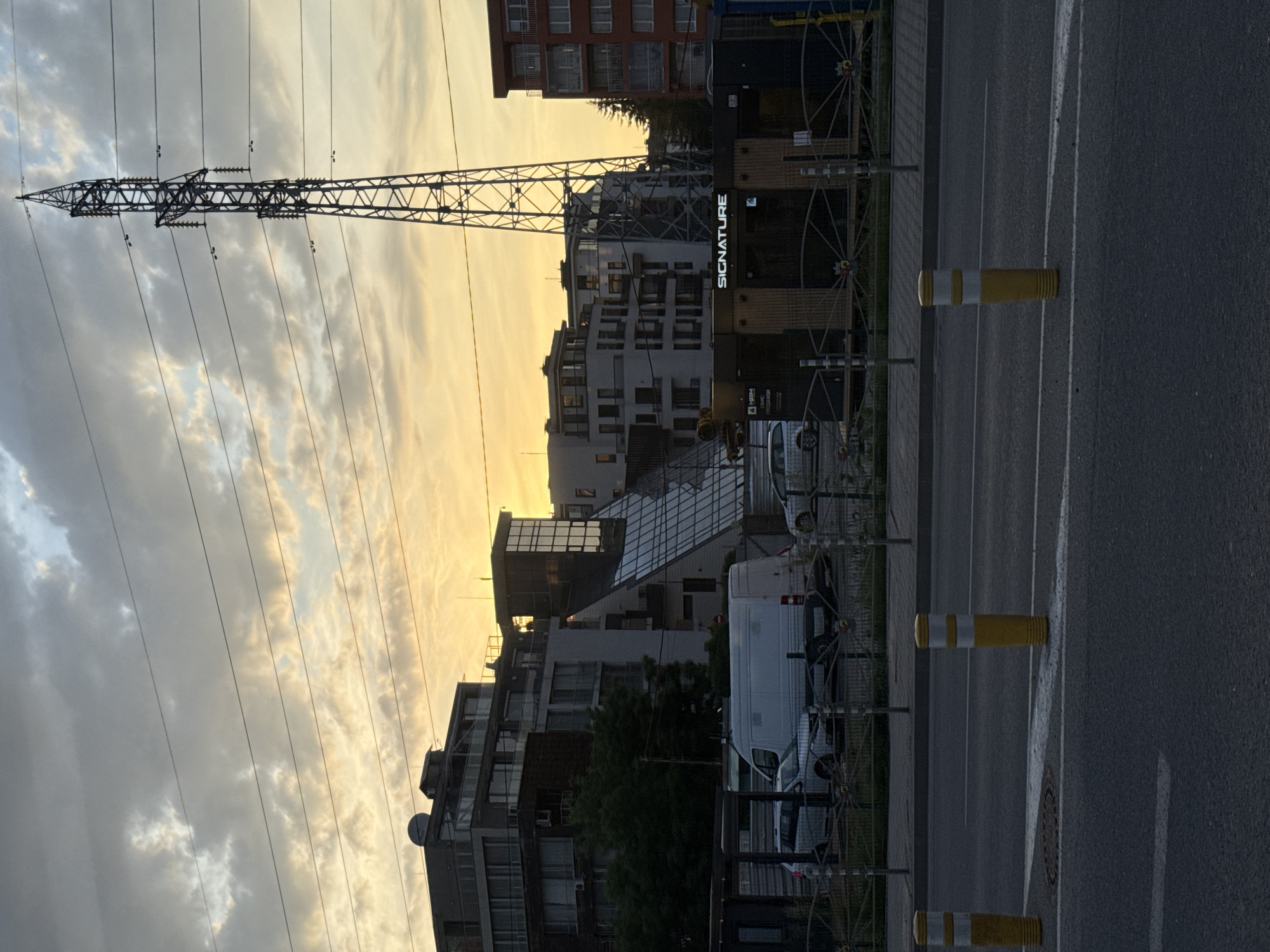 The image shows an urban landscape in Sofia, Bulgaria, at sunset. A high-voltage power line tower dominates the left side of the frame against a cloudy sky with a warm, golden hue. In the background, there are several buildings, including modern residential structures with glass elements, and a building with the word "SIGNATURE" prominently displayed. A row of yellow and white striped bollards lines the edge of a street, suggesting a parking area. White vans are parked along the side of the road behind a decorative metal fence. The asphalt street occupies the right side of the frame, with painted lane markings and a manhole cover visible. The overall scene conveys a blend of urban infrastructure, residential architecture, and transportation elements.