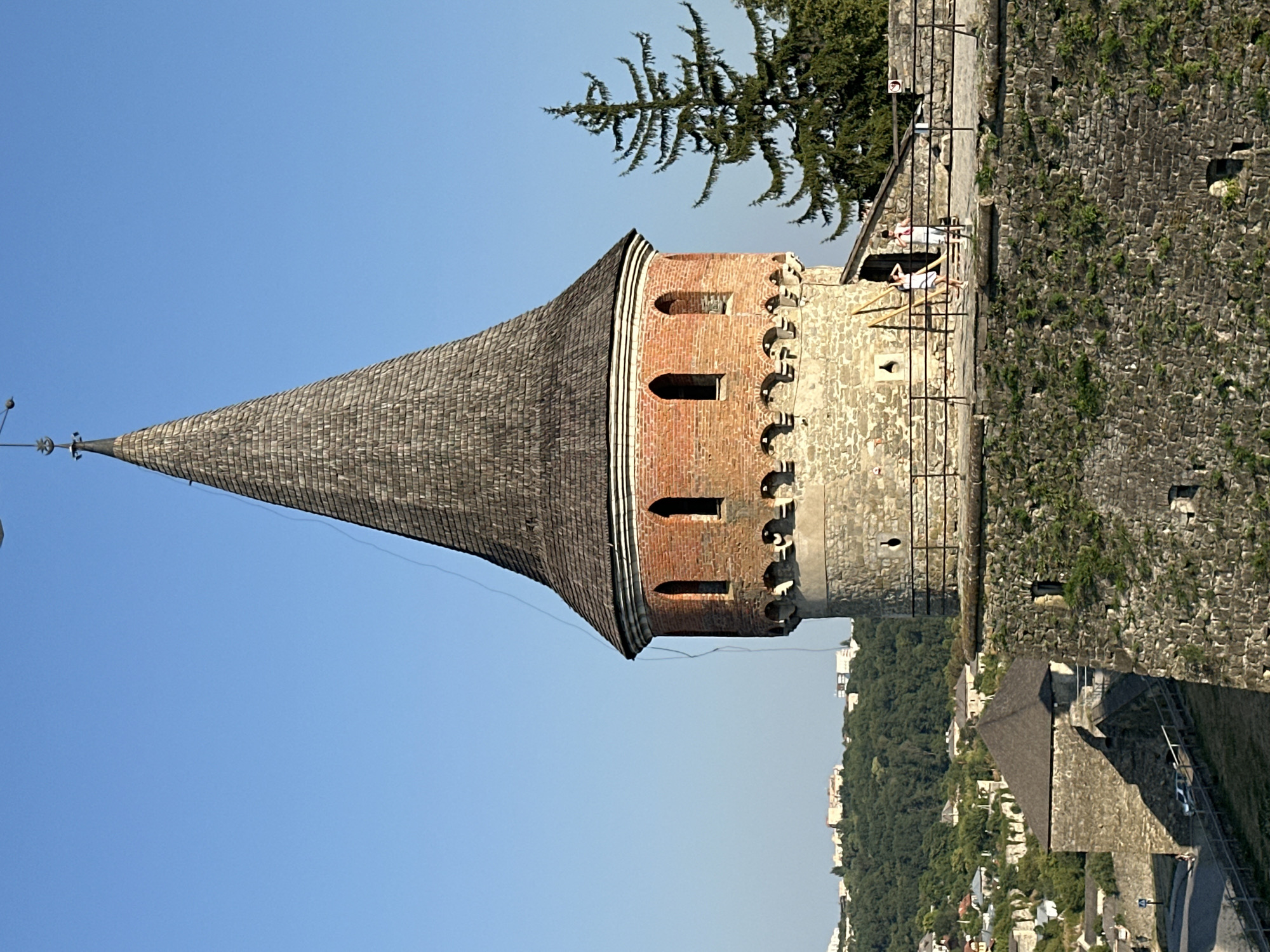 The image captures a close-up view of a medieval tower, a prominent feature of the Kamianets-Podilskyi Castle in Ukraine. The tower boasts a conical, wooden shingle roof, topped with a weather vane. Below the roof, the tower is constructed of red brick with arched window openings and a decorative crenellated parapet. The lower portion of the tower transitions to rough-hewn stone. A stone walkway, bordered by a metal railing, extends from the tower, with two individuals visible walking on it. In the background, a lush green landscape dotted with buildings and a tall evergreen tree suggests a settlement on a hillside. The sky is a clear, bright blue.