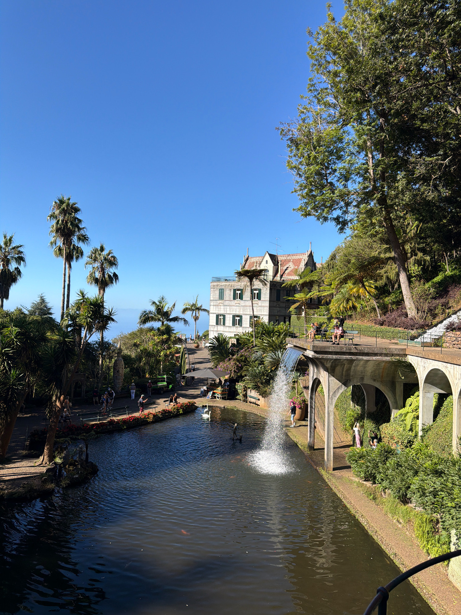 The image depicts a scenic view of the Monte Palace Tropical Garden in Funchal, Portugal. In the foreground, a tranquil pond with dark, rippling water reflects the surrounding greenery and sky. Two white swans are visible on the surface of the pond. Several koi fish can be seen near the surface of the pond. A pathway lined with red flowers runs along the edge of the pond, with several people strolling along it.
In the mid-ground, a multi-story building with white walls and a red-tiled roof stands prominently. Its architecture suggests a European style, possibly a historic villa or a garden pavilion. A path leads up to this building, and more people are seen walking and sitting on benches. A lush garden with a variety of trees and plants, including palm trees, surrounds the building and pathways.
To the right of the frame, a stone archway structure is visible, with a waterfall cascading from its upper level into the pond. The arches are supported by thick pillars, and the structure is covered in dense green foliage. A walkway runs along the top of the archway, providing a vantage point over the garden and pond. A few individuals are present on this walkway, observing the scenery. The background reveals a hazy blue horizon, suggesting the presence of the ocean. The sky above is a clear, bright blue. The overall atmosphere is serene and picturesque, characteristic of a well-maintained botanical garden on a sunny day.