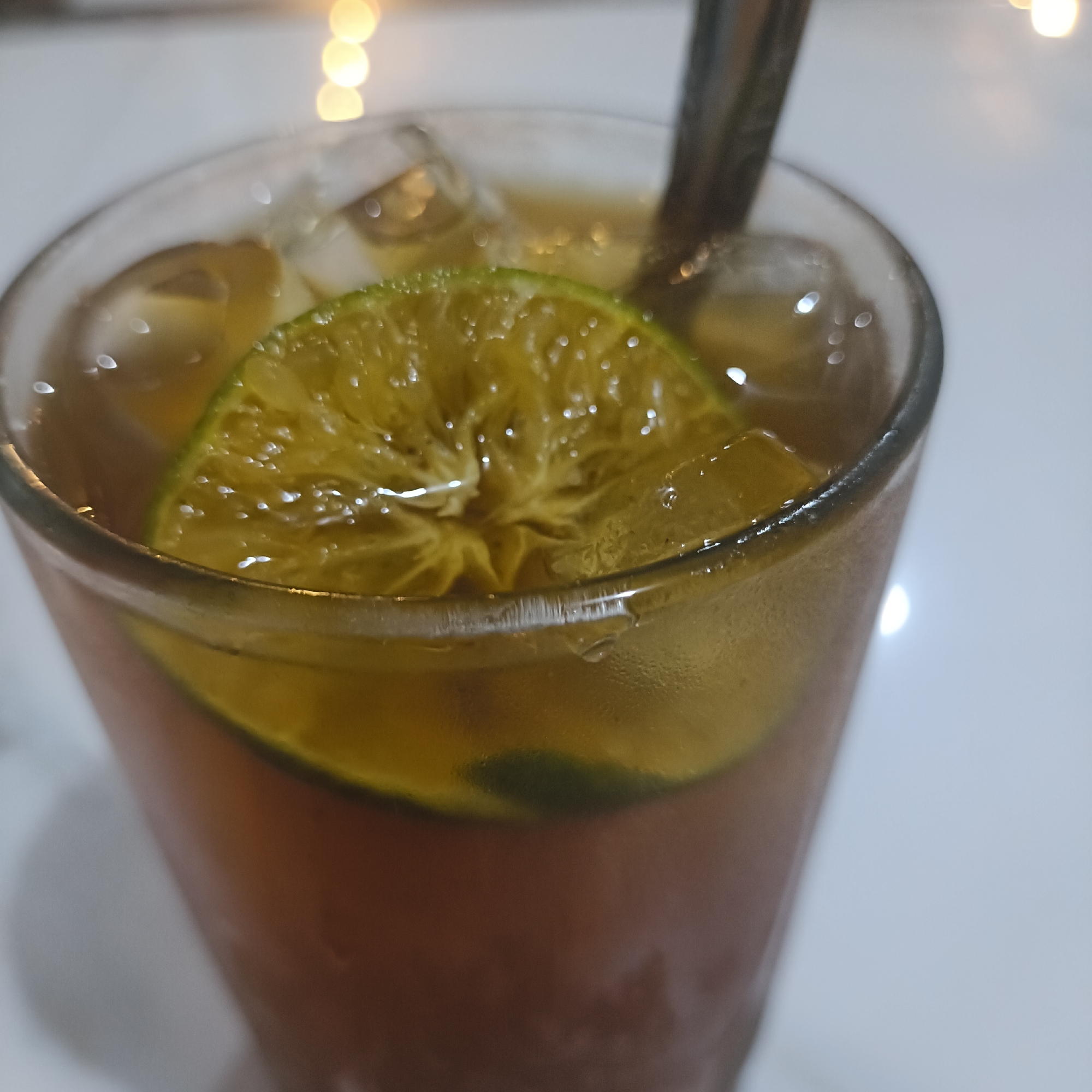 A glass of iced tea with a lime slice and a spoon is shown close-up. Condensation is visible on the glass. The background is blurred, showing what appears to be a table surface and distant bokeh lights. The location is implied to be in Cangkring, Indonesia.