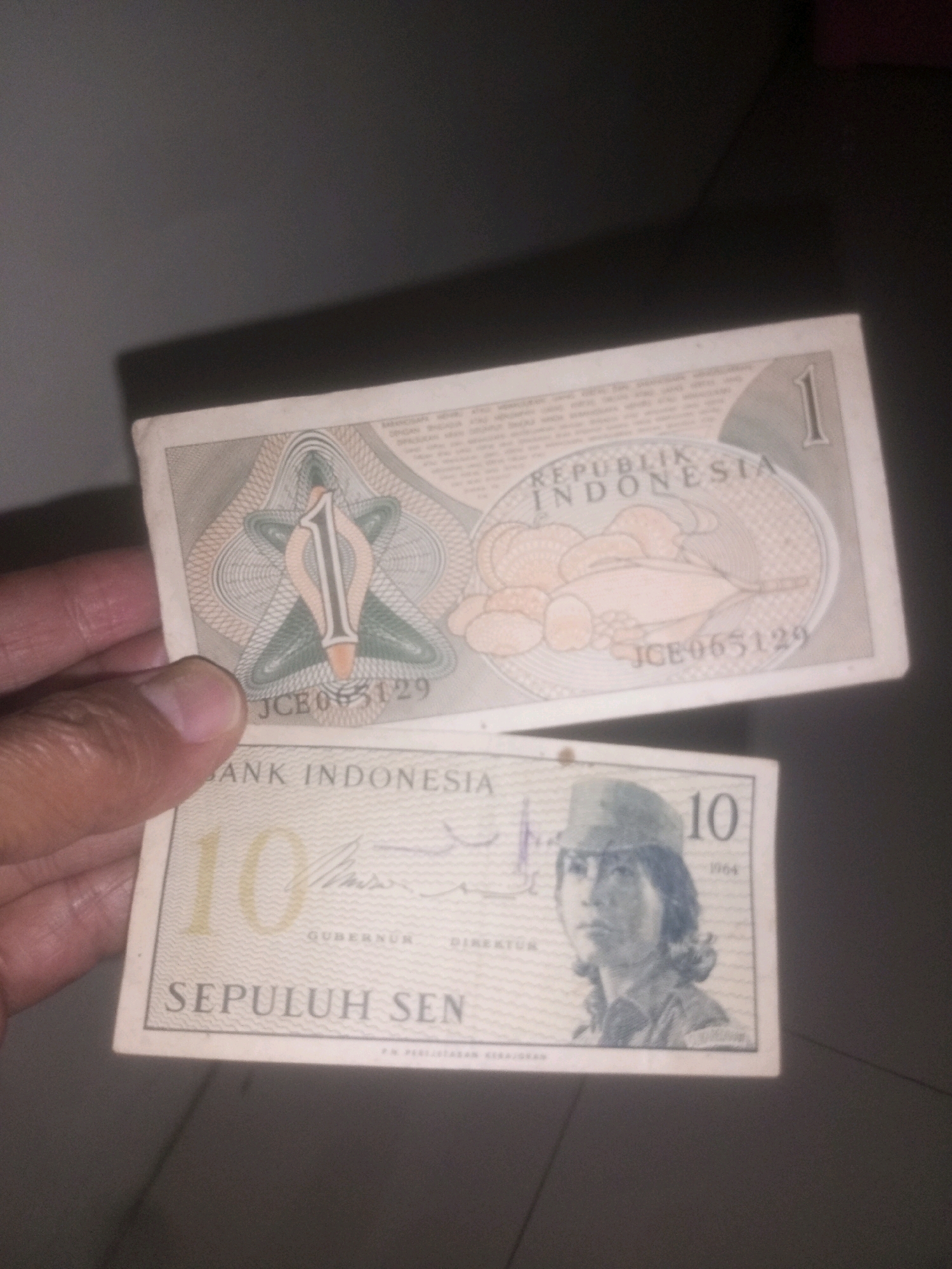 A pair of Indonesian banknotes are held in a person's hand against a dark, textured background. The top banknote is light beige with a circular emblem featuring the number "1" and the text "REPUBLIK INDONESIA". Below this emblem, the serial number "JCE065129" is visible. The bottom banknote is also light beige and features the number "10" and the text "BANK INDONESIA". A portrait of a person wearing a hat and a shirt is depicted on the right side of this banknote, along with the text "SEPULUH SEN" and the year "1964". The background is indistinct but appears to be a wall or dark surface. The image is taken from a slightly downward angle. There is no indication of the city of Kediri City, Indonesia, in the image.