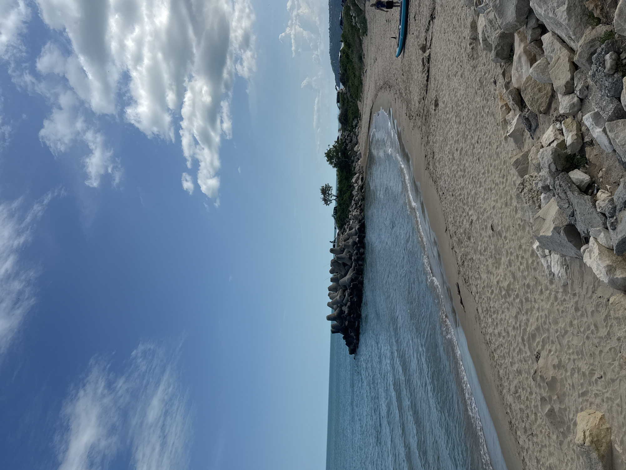 The image depicts a beach in Varna, Bulgaria. The scene is divided vertically, with the sky and clouds on the left and the coastline on the right. The beach consists of sand, rocks, and a concrete breakwater extending into the sea. Small waves are visible along the shore. Vegetation and possibly some parked cars line the top edge of the beach. At the upper right of the beach, two individuals and a blue paddleboard can be seen.