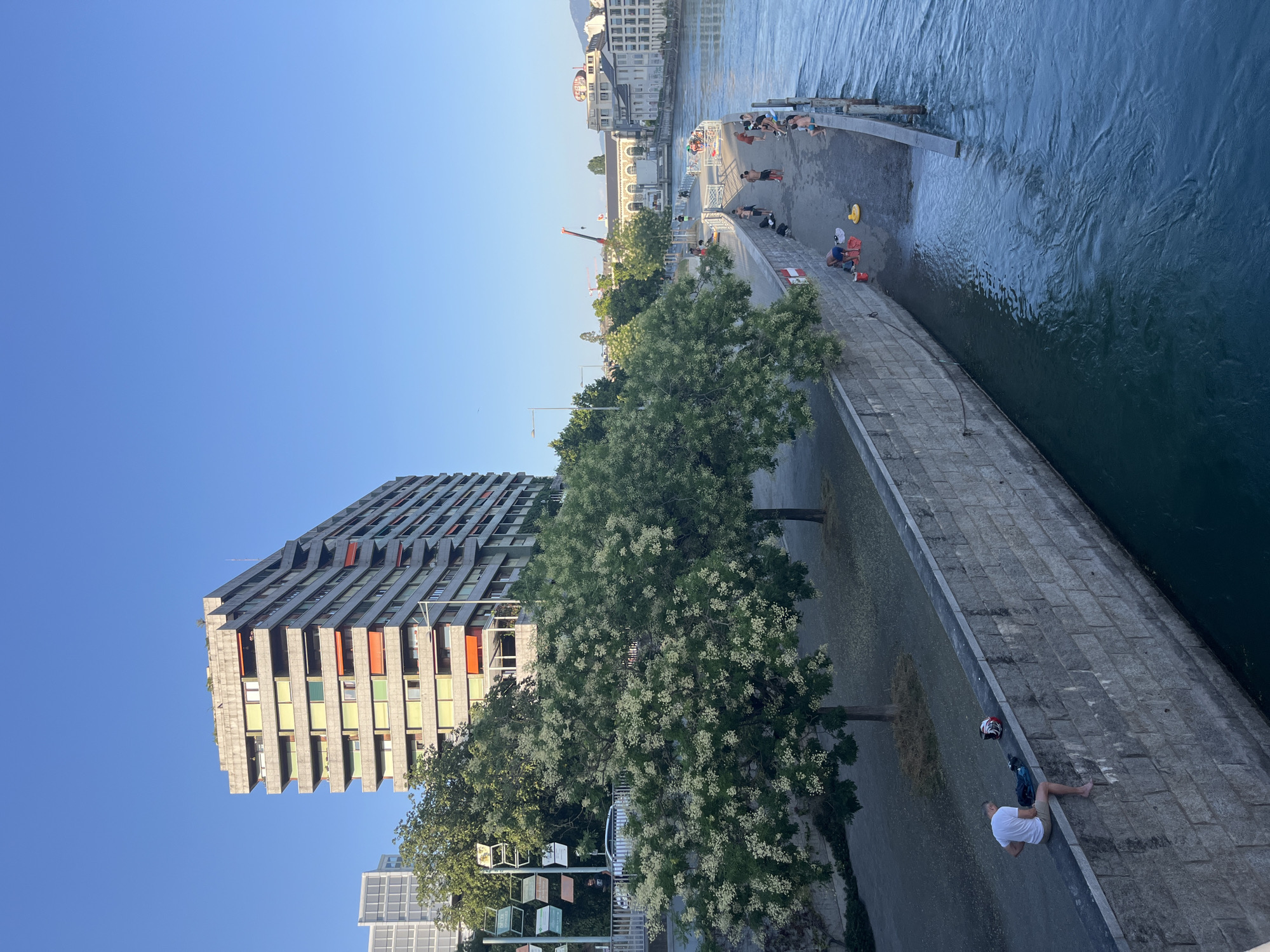 This image depicts a sunny day along the Rhone River in Geneva, Switzerland, near the Pont des Bergues. On the left, a large, multi-story apartment building with a gray facade and numerous balconies is visible, some adorned with orange awnings. Adjacent to the building are lush green trees with white blossoms, shading a pedestrian walkway.
The riverbank features a stone embankment. Further along the embankment, a paved area is populated with people, many of whom are dressed in swimwear, suggesting recreational activity. Some individuals are sitting, while others appear to be interacting or preparing to enter the water. A man in a white t-shirt and shorts is seated on the stone wall, his bare feet dangling over the water, with a helmet and bag beside him. The river itself is a vibrant blue-green, with visible ripples indicating flow. In the background, other buildings line the opposite bank of the river, and a crane is visible further in the distance, hinting at ongoing construction or maintenance in the city. The sky is a clear, bright blue.