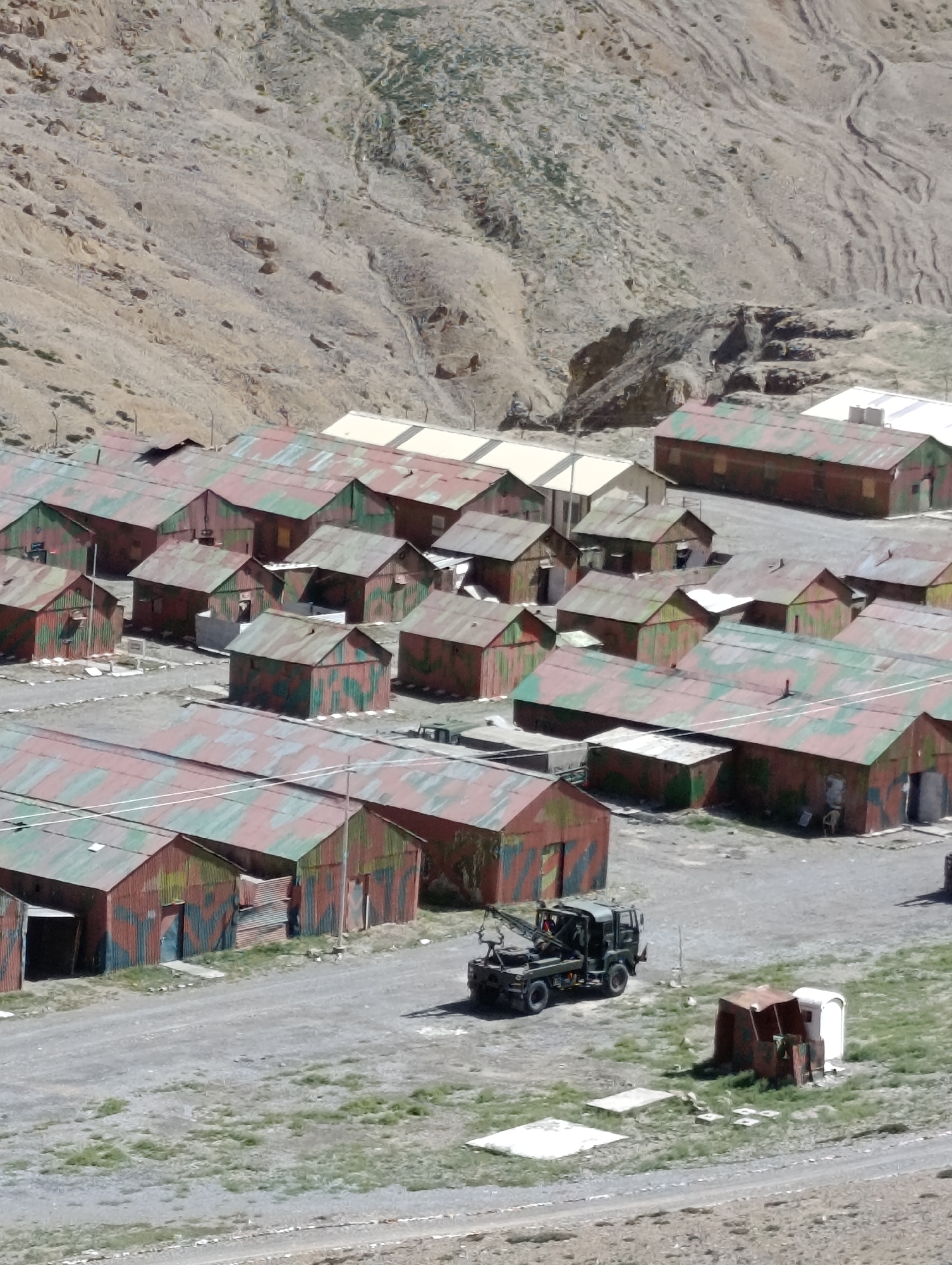 A high-angle, eye-level view shows a military encampment on a hillside in an arid, mountainous region of India. The encampment consists of numerous small, corrugated metal buildings with camouflaged roofs in red and green. The buildings are arranged in rows with gravel pathways between them. In the center of the frame, a dark green military truck with a crane is parked on a gravel road. To the right of the truck, a small, open-sided shelter with a white portable toilet is visible. The background is dominated by a steep, barren, rocky hillside with sparse vegetation and evidence of water erosion. The sky is not visible in the frame.