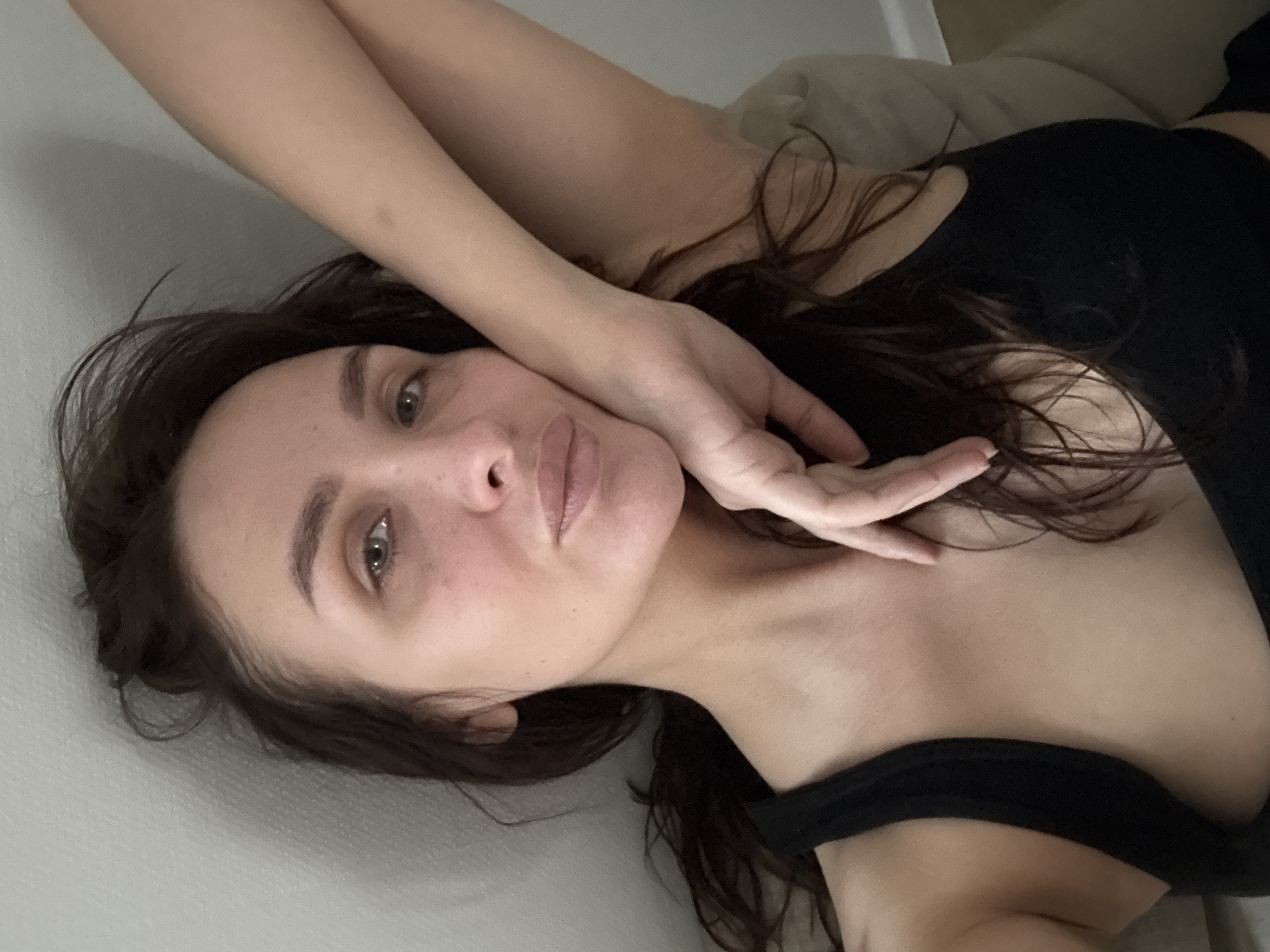 The image presents a close-up, upward-angled view of an adult female, likely a selfie. The individual has long, dark brown hair and light-colored eyes, appearing to be of European descent with a light skin tone that shows minor textural variations and freckles/moles. She is wearing a black tank top or sports bra. Her left hand rests against her left cheek and chin, with fingers slightly spread. Her right arm is bent and partially visible in the upper right section of the frame, suggesting a reclined or lying posture. The subject is looking directly into the camera with a neutral facial expression and slightly parted lips. The background consists of a plain, light-colored wall to the upper left and behind her head, transitioning to a soft, light-toned object, possibly a cushion or part of a bed, to her right and behind her shoulder. The visual content of the image does not provide specific details to confirm a location within the city of Chaikovsky, Russia.