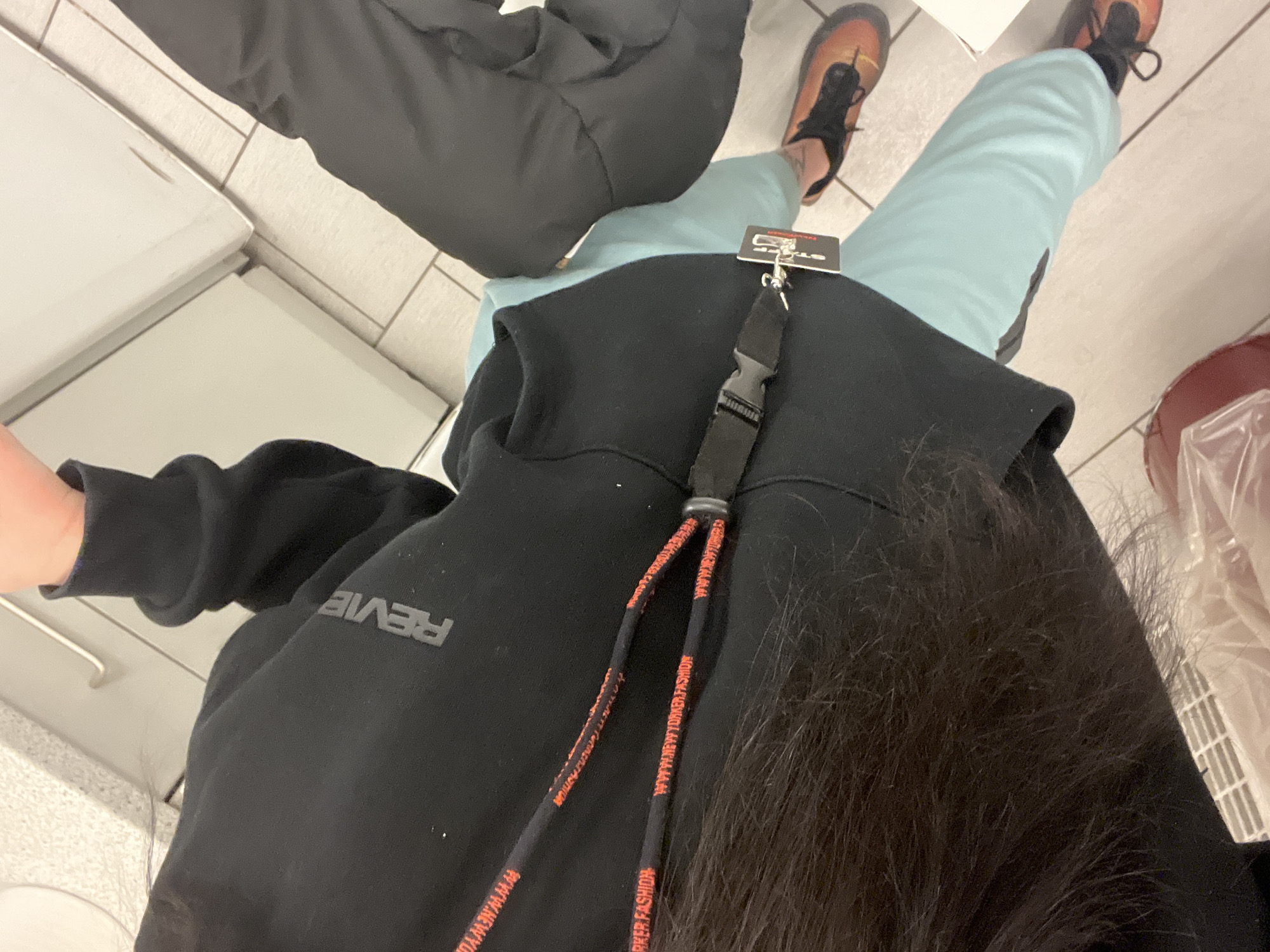 An overhead shot depicts an indoor commercial environment, likely a retail store or shopping mall in Sofia, Bulgaria. The primary subject, viewed from the chest down, wears a black long-sleeved top with the word "REVIEW" visible in grey on the right sleeve. A black lanyard with repeating orange text "WWW.NEWYORKER.FASHION" and "NEW YORKER FASHION" is worn around the neck, secured by a black plastic buckle, from which a white tag and a set of keys are suspended. The individual wears light blue pants and black and orange athletic shoes. Dark hair is partially visible on the left side of the frame. To the right, the lower torso and legs of a second individual are partially visible, wearing dark grey or black pants. The floor consists of light-colored rectangular tiles. In the background, a white counter or fixture is visible. A red container with a clear plastic bag is partially seen in the bottom left, and a white rectangular object is in the bottom right.