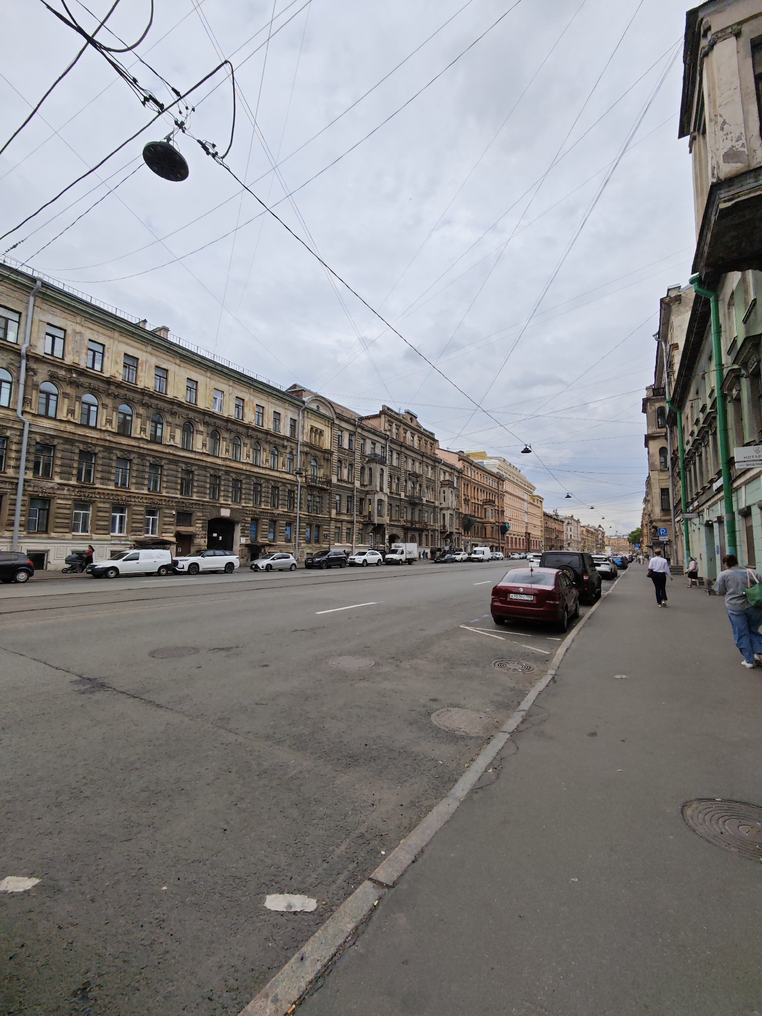 The image shows a street scene in Saint Petersburg, Russia, with a wide asphalt road in the foreground and buildings lining both sides. The sky is overcast. On the left side of the road, there are several multi-story buildings with ornate facades and numerous windows. A row of parked cars and vans, including white commercial vehicles and passenger cars, are visible along the sidewalk. Some people are walking on the sidewalk.
On the right side of the road, there are also buildings, some of which appear older and have peeling paint. A sidewalk runs along these buildings, with pedestrians walking. Several cars are parked along the street. Traffic lights or street lamps hang over the road, supported by a complex network of wires. The road surface shows some signs of wear and tear, with manhole covers and patches visible. The overall impression is of a busy urban street in an older part of the city.