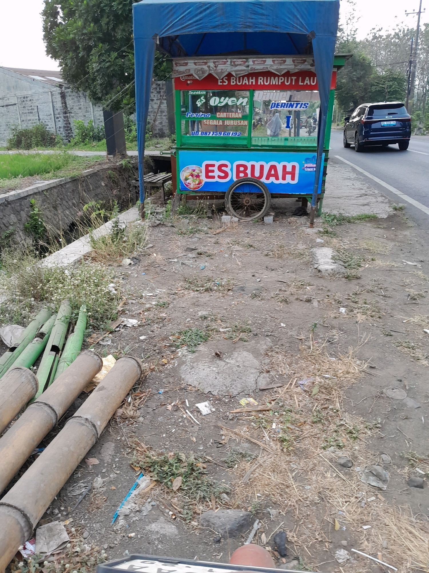 This is a medium-shot, eye-level, outdoor photograph taken on a slightly overcast day, showing a street-side food cart. The cart, predominantly blue and green, is positioned on the right side of the image, partially obscuring the road. It is adorned with signage, including "ES BUAH RUMPUT LAU" and "ES BUAH". A spinning wheel is attached to the cart.
In the background, a dark blue SUV is driving away on the road. To the left of the cart is a drainage ditch with a stone embankment. Beyond the ditch, there is a field of green grass and a textured wall. In the foreground, a pile of large bamboo poles lies on the ground, along with scattered debris including dry grass, leaves, and plastic waste. The ground is a mixture of dirt and gravel.
The scene appears to be in the village of Pakisaji, Malang Regency, East Java, Indonesia, as indicated by the presence of "Kabupaten Malang" on the vehicle's license plate, which is partially visible. No people are clearly visible in the frame. The overall impression is one of a roadside scene with a food vendor, a vehicle on the road, and the natural and man-made elements of the environment.