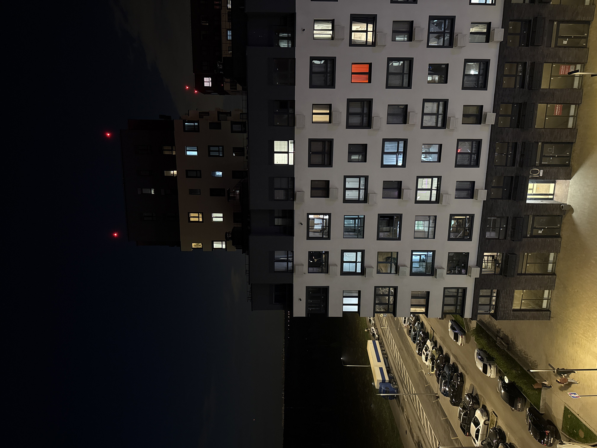 View of buildings and a street scene at night in Troitsk, Russia. Two tall residential buildings occupy much of the frame, with numerous lit and unlit windows; one building is predominantly white, the other is dark. Red lights are visible atop one of the darker buildings. Below, a street contains parked cars and a large truck. Two people are seen walking on a sidewalk on the right side of the frame. The left side shows a dark sky and horizon line.