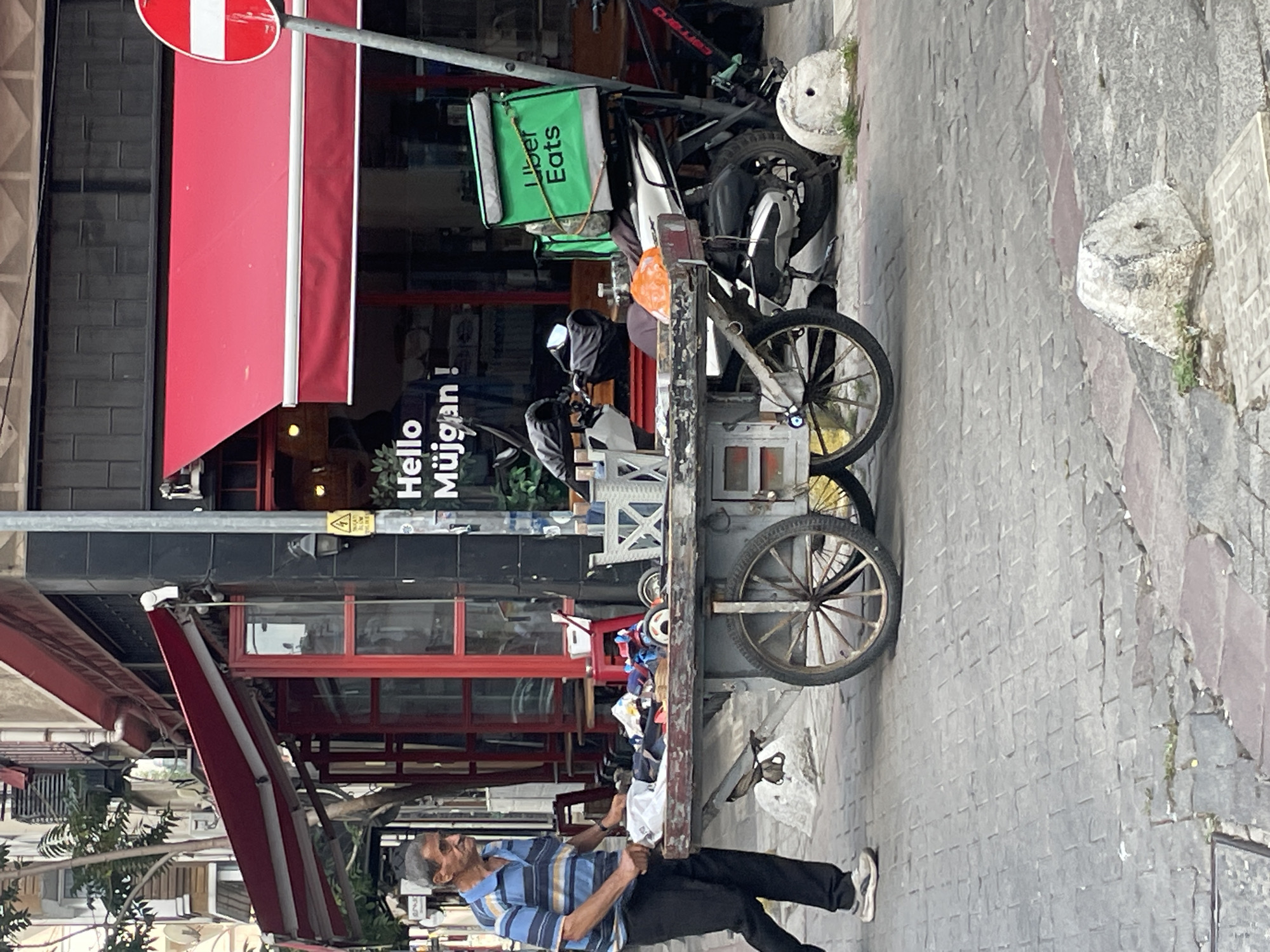 A man walks pushing a cart on a cobblestone street in Kadıköy, Turkey. The man is wearing a striped blue and white shirt, dark pants, and white sneakers. The cart is made of wood and metal and is laden with various items, including bags, containers, and what appears to be food. In the background, there is a building with a red awning and windows. A red and white circular "do not enter" sign is visible on a pole. A motorcycle with a green Uber Eats bag is parked nearby. The scene suggests a street market or a vendor's stall.