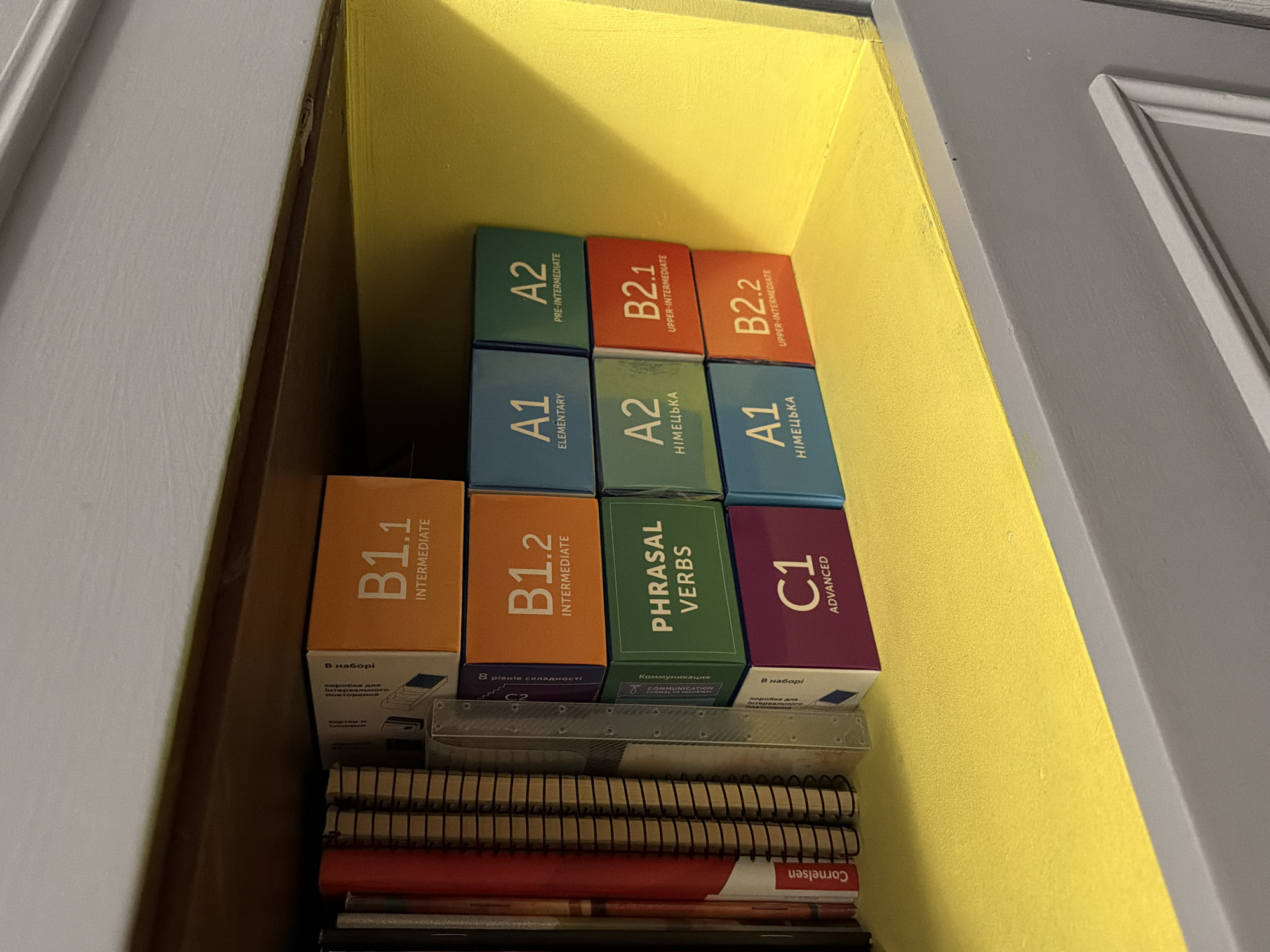 A close-up shot shows a shelf filled with various items. At the top are several language learning boxes stacked in a pyramid formation. These boxes display language levels such as A1 (Elementary), A2 (Pre-Intermediate), B1.1 (Intermediate), B1.2 (Intermediate), B2.1 (Upper-Intermediate), B2.2 (Upper-Intermediate), and C1 (Advanced). One box is labeled "Phrasal Verbs." Below these boxes are notebooks and what appears to be a thin book or folder labeled "Cornelsen." The background is a yellow interior of the shelf, with the edges defined by grey door frames.