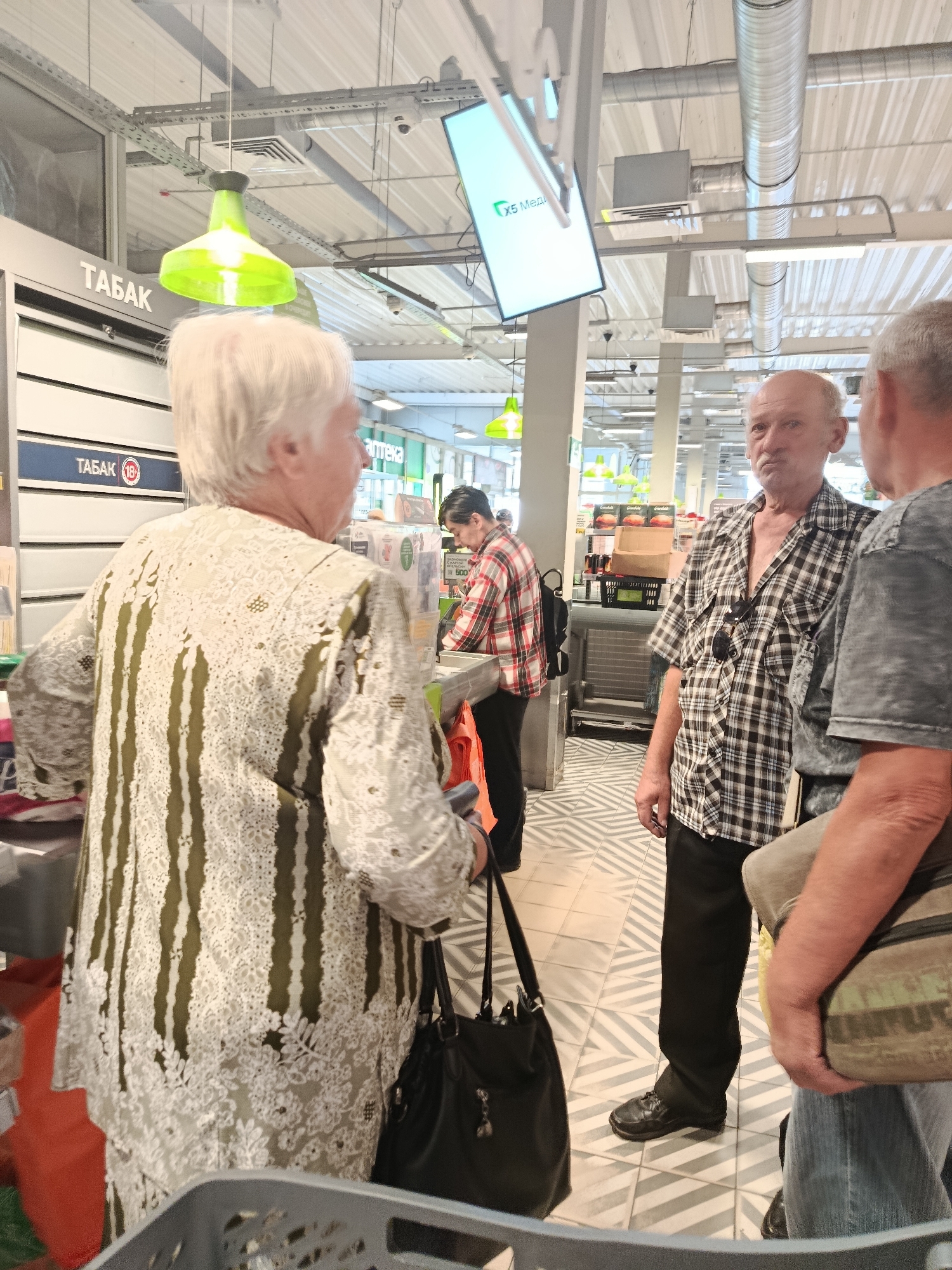 A medium shot captures an interior scene within what appears to be a retail environment, possibly a supermarket or convenience store, in Orekhovo-Zuyevo, Russia. Three individuals are visible. In the foreground, an older woman with white hair, facing away from the camera, is wearing a patterned blouse with white lace overlay and green stripes. She is holding a black handbag. To her right, a bald, middle-aged man in a black and white checkered shirt and dark pants looks towards the camera with a serious expression. He is wearing sunglasses on the collar of his shirt and has a bag slung over his right shoulder. Partially visible on the far right is another person, whose arm and shoulder are in frame, wearing a gray t-shirt and holding a camouflage bag. In the background, another individual in a red and white plaid shirt is seen interacting with a checkout counter. The environment features overhead lighting, ceiling fixtures, and various signage. One sign reads "ТАБАК" (Tobacco) with a circled "18" indicating an age restriction. Another sign displays "X5 Медиа" (X5 Media), suggesting a connection to a retail chain. The flooring is a patterned mosaic of light and dark gray diagonal lines. The overall impression is a candid moment captured in a public retail space.