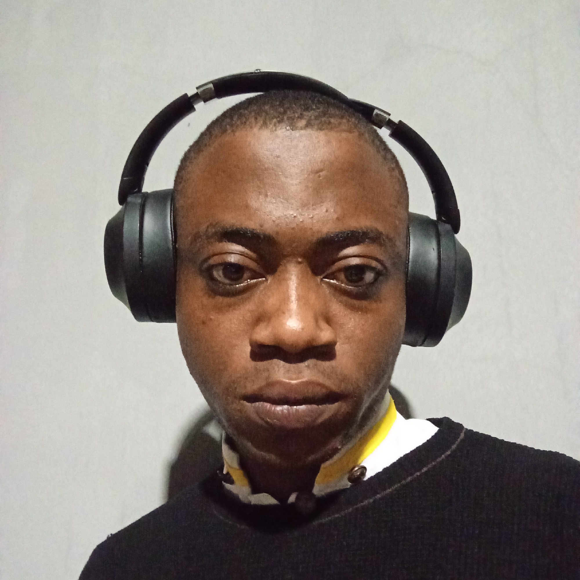A medium shot of a young Black male wearing large, black over-ear headphones, a black sweater, and a collared shirt with yellow and white accents. The background is a plain, light gray wall. The man's eyes are open and he is looking directly at the camera. The lighting is somewhat dim, casting shadows on his face and the background. The image appears to be a selfie taken indoors in a residential setting, likely in the city of Ibadan, Nigeria, given the context provided. There is no discernible activity or interaction taking place; the focus is solely on the subject.