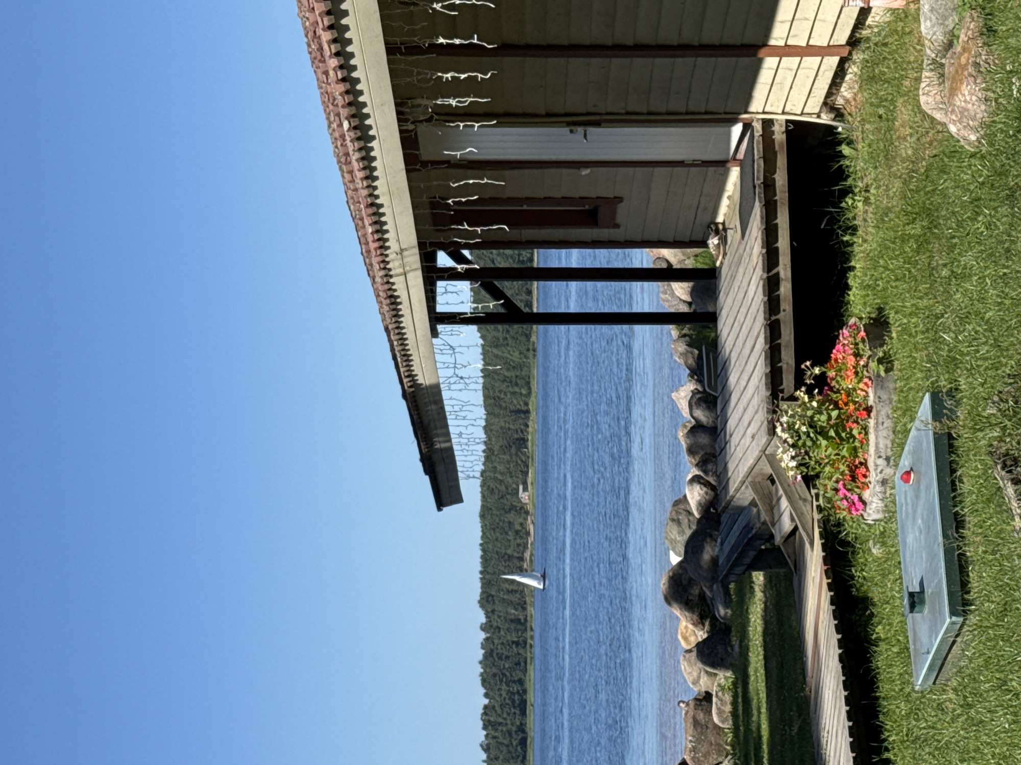A wooden cabin with a tiled roof and string lights is situated on the grassy bank of a lake. A wooden deck extends from the cabin towards the water. In the foreground, a metal access panel sits in the grass. The lake is calm, with a sailboat visible in the distance and a dense forest lining the opposite shore. The sky is clear and blue. The scene is located in Ozerki (Krasnodolinskaya), Russia.