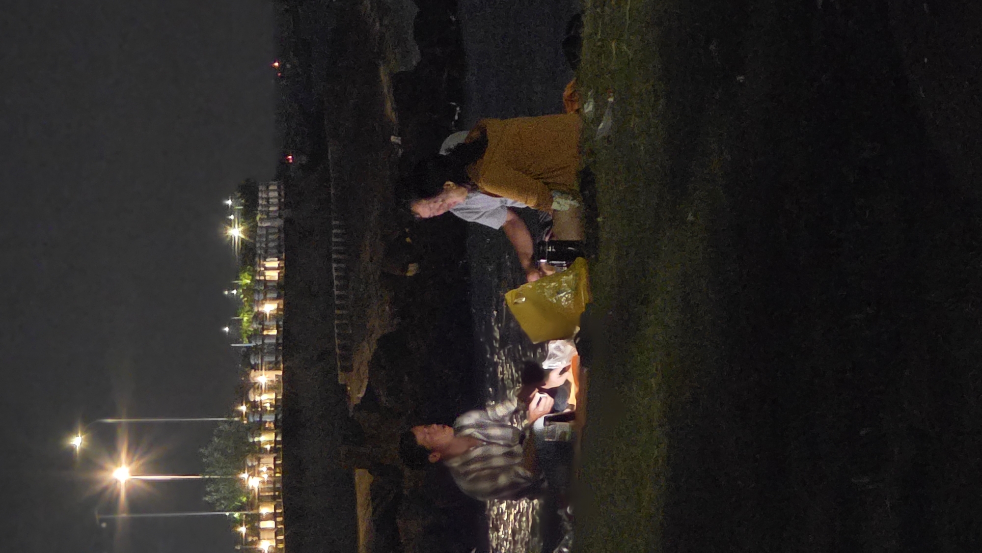 At night in Bắc Nha Trang, Vietnam, an eye-level shot captures two individuals along the seawall. One person in a checkered shirt and dark jeans is seated at the edge, while another in an orange shirt is also seated, with a yellow bag at their feet. The focus is on the people and their interaction with the seawall. In the background, the cityscape lights illuminate the scene, contrasting with the dark sky.