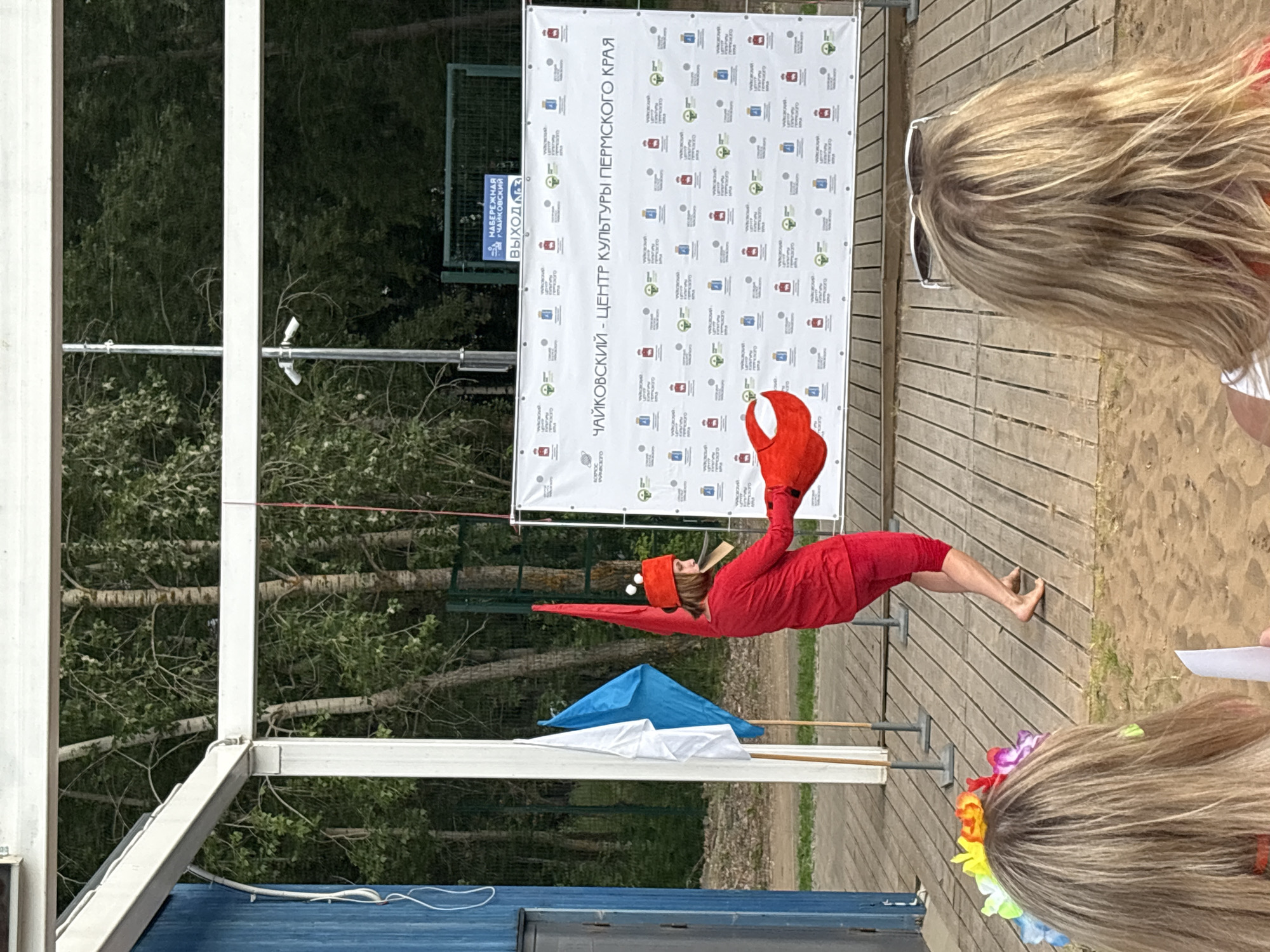 This media file depicts an outdoor event on a sunny day, likely in Chaikovsky, Russia. In the foreground, the back of a person's head with blonde hair adorned with a colorful lei is visible. A larger banner in the background displays text in Russian, which reads "ЧАЙКОВСКИЙ - ЦЕНТР КУЛЬТУРЫ ПЕРМСКОГО КРАЯ" (Chaikovsky - Center of Culture of the Perm Krai) and "НАБЕРЕЖНАЯ Г. ЧАЙКОВСКИЙ" (Embankment of Chaikovsky City). The banner also shows various logos.
The central focus of the image is a performer dressed in a vibrant red costume, resembling a crab, with large red claws. The performer is in a dynamic pose, appearing to be suspended in mid-air or performing a contortionist act. They are barefoot and wearing a matching red cap. Two colorful umbrellas, one blue and one white, are positioned behind the performer, possibly as props or for shade. The setting appears to be a wooden boardwalk or stage area next to a sandy patch, with trees and foliage visible in the background, suggesting a park or recreational area. The overall atmosphere seems festive and engaging, likely a cultural event or performance.